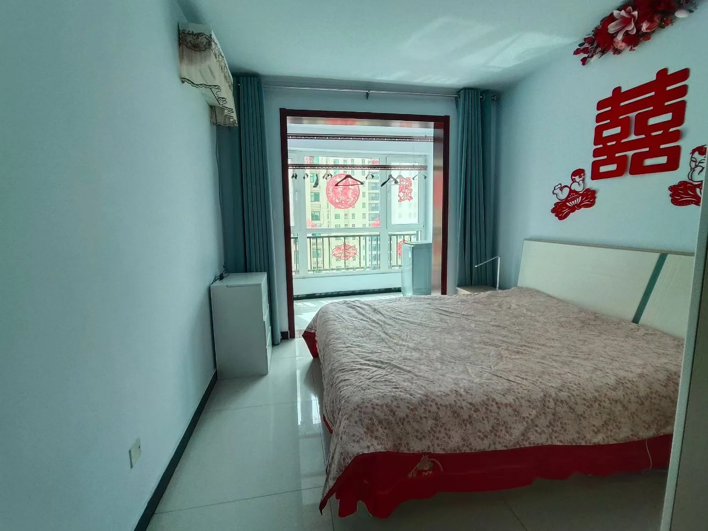 卧室场景，墙面装饰有醒目的红色中式图案，床铺整洁，采光良好，整体呈现温馨而具有传统装饰风格的居住空间。