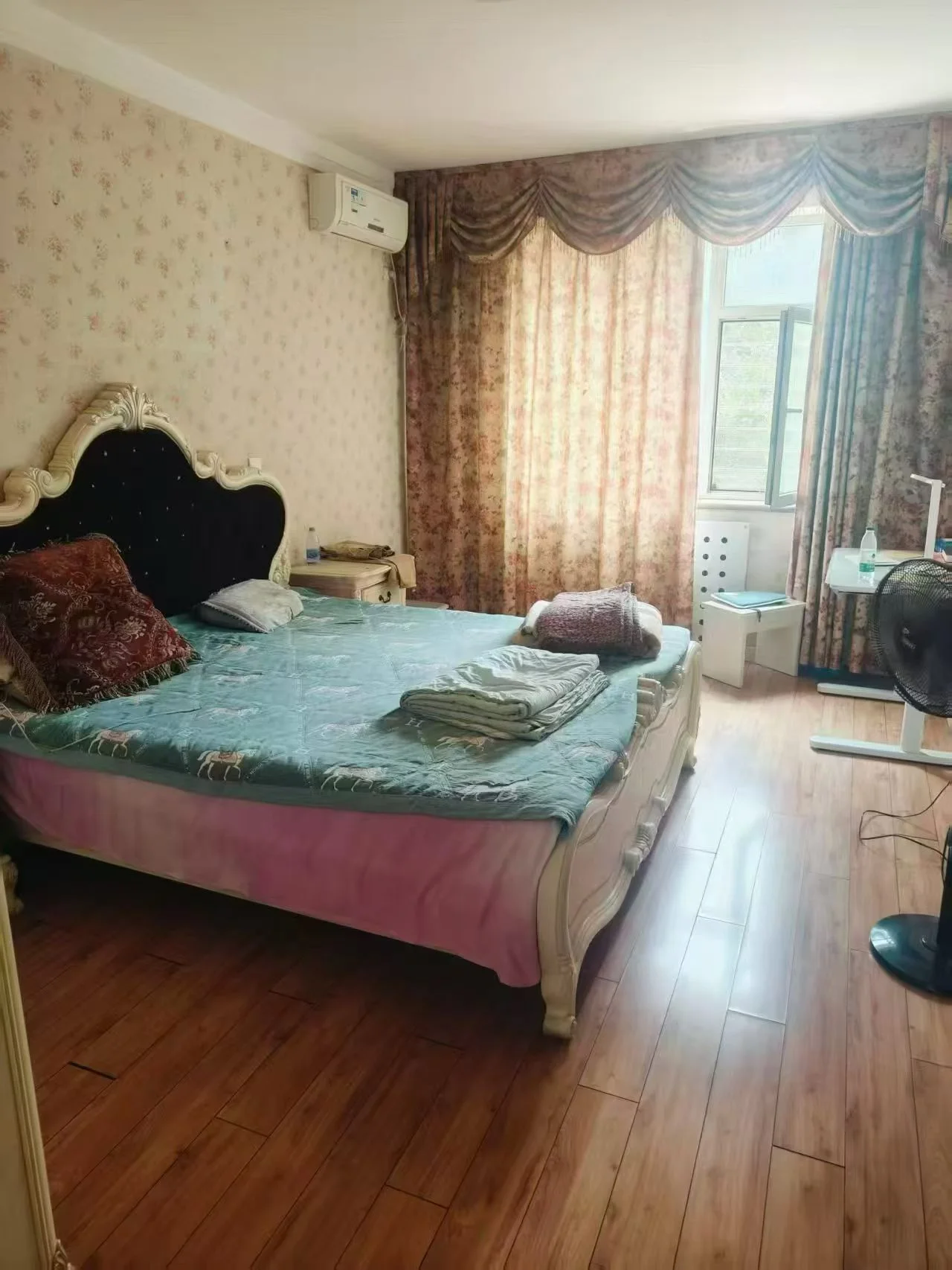 卧室场景，床铺铺设了蓝绿色床单，配有深色床头板和窗帘。采光良好，整体呈现温馨、朴素的居住氛围。