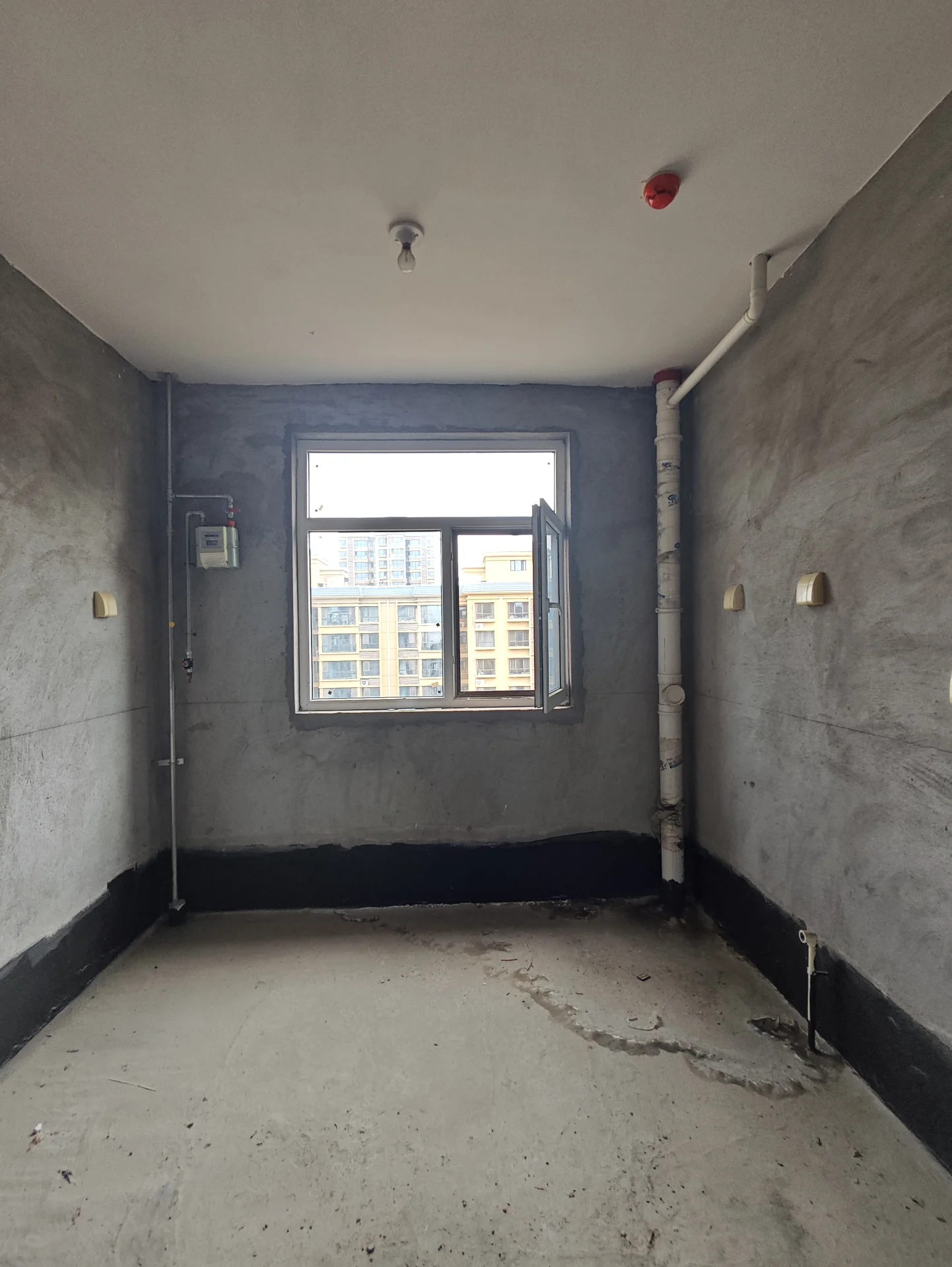 混凝土地面和墙面的房间，似乎正在施工或翻新中。房间有窗户和沿着墙壁的管道，给人一种工业或未完成的感觉。氛围像是施工或翻新区域，有管道和房间未完成的外观。