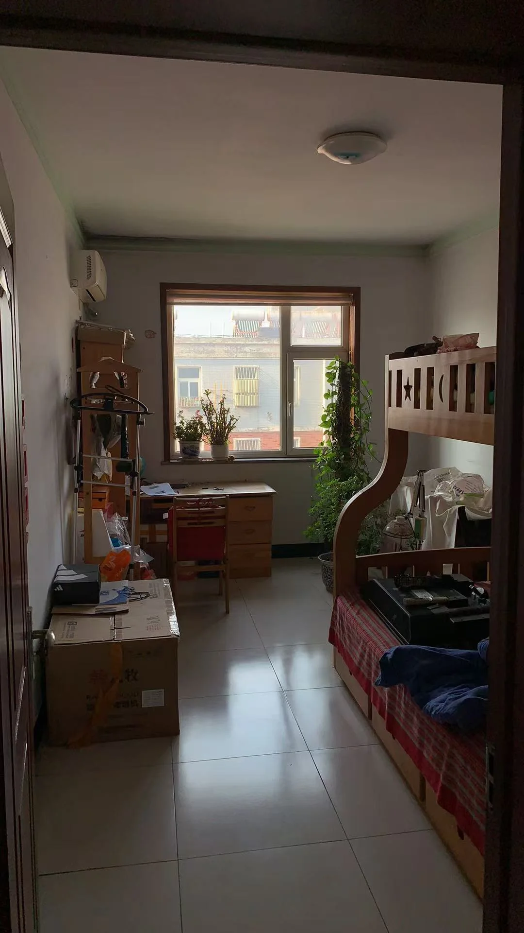 一个房间的内部场景，地面铺设米色瓷砖，墙面为浅色调，配有窗户采光良好。空间内摆放着木质储物柜和一些家具，整体呈现一个朴素、功能性强的居住环境。