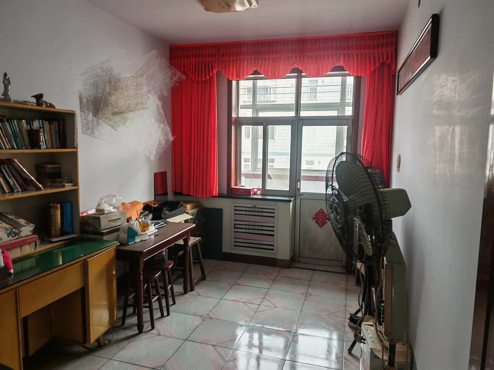 杂乱房间，有窗户和风扇，地板上有书，椅子、餐桌和书桌，窗户有红色窗帘。