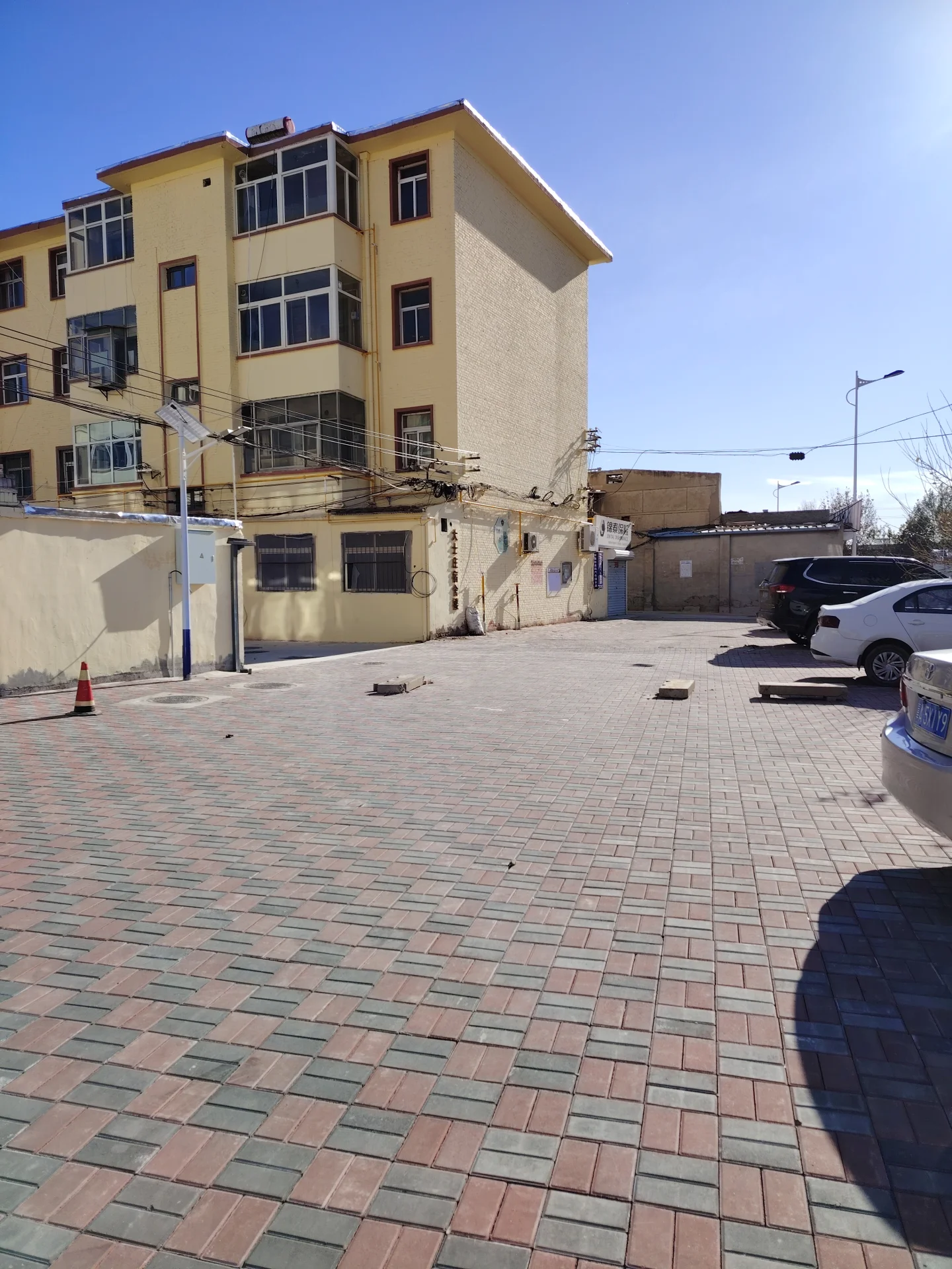 一栋米黄色多层住宅的外部立面，建筑结构简洁，配有规则的窗户。建筑前方是铺设了红棕色砖的地面，右侧停放着几辆汽车，整体呈现社区或住宅区的外部环境氛围。