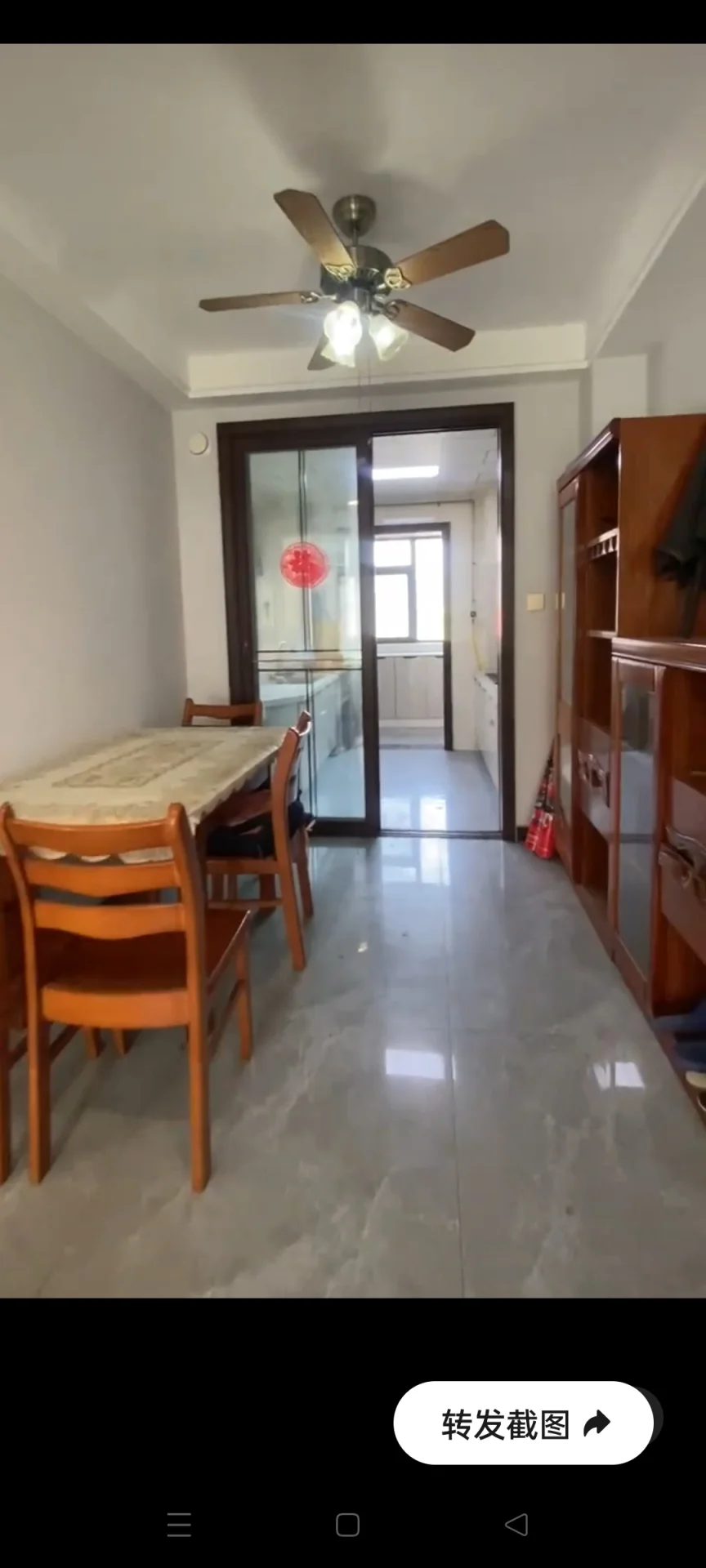 一个宽敞的室内空间，地面铺设了浅色大理石纹理的地板，配有木质餐桌和椅子。房间内安装了吊扇，光线充足，整体呈现温馨、朴实的居住氛围。