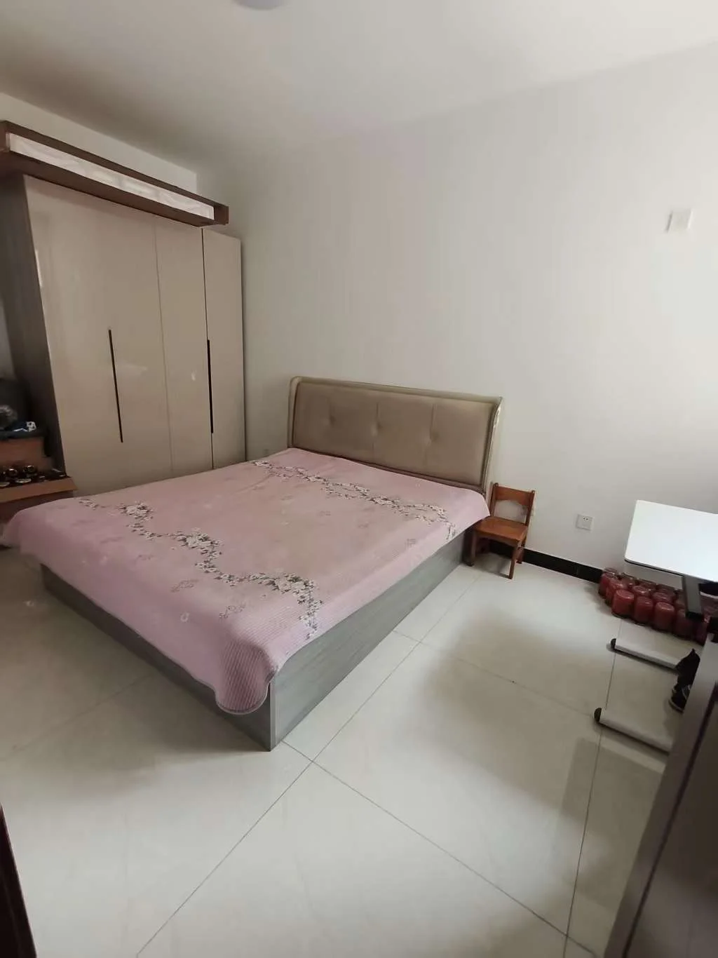 卧室场景，床铺铺着粉色花纹床单，床头有木质床架和衣柜，地面铺设白色瓷砖，整体装修风格简洁明亮，采光良好，营造出整洁舒适的居住氛围。