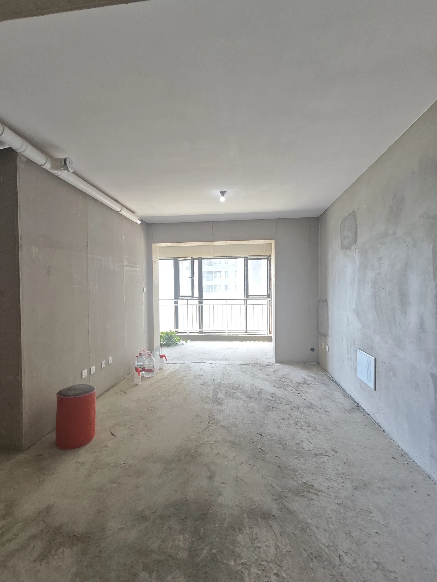 空旷房间，地面为混凝土，天花板为白色，房间正在进行施工或翻新，散落着各种施工材料如红色桶和红色凳子。房间内有窗户，可能是在施工过程中的一部分。整体场景给人一种正在改造或改善的空间的印象。