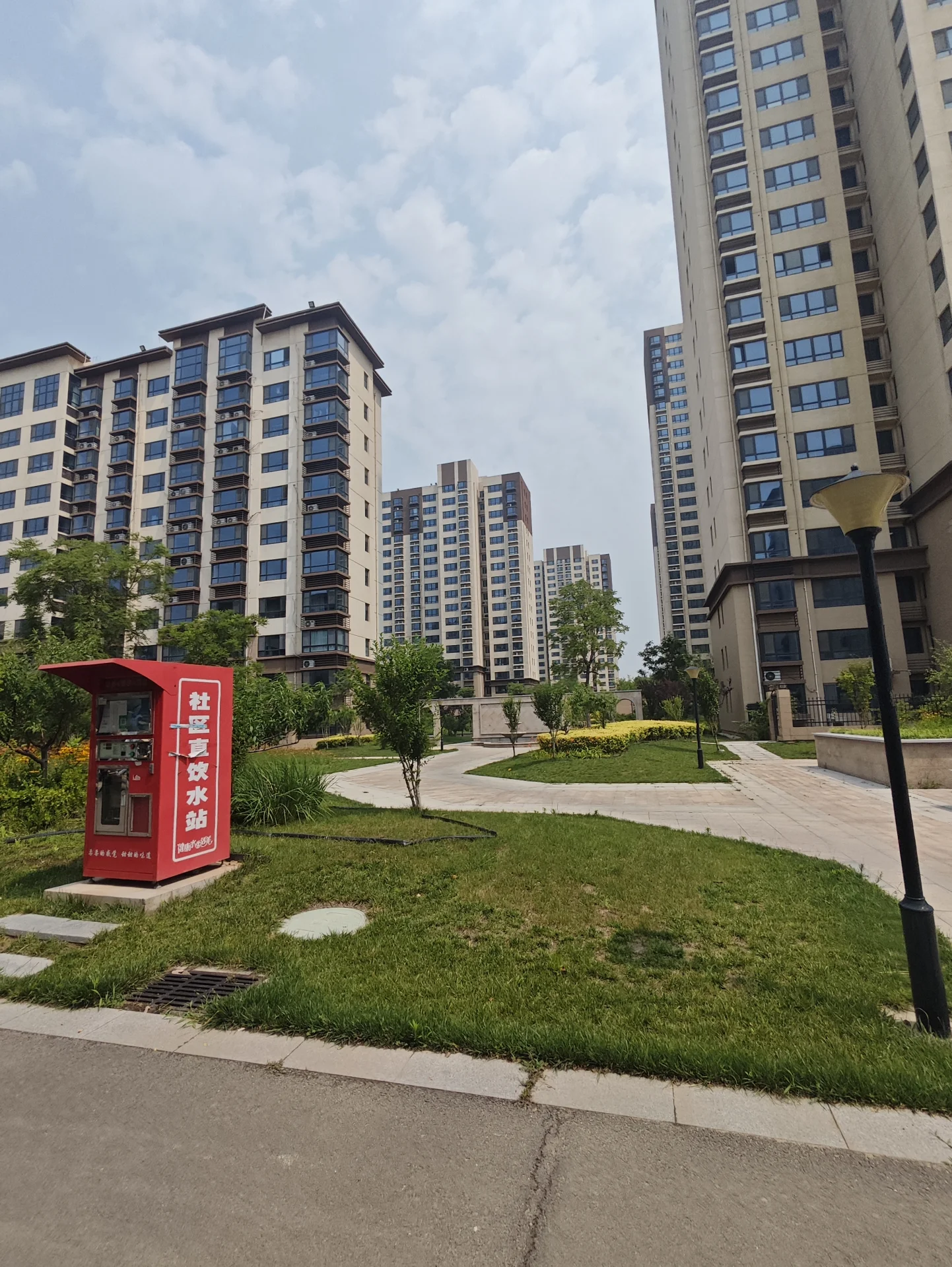 多栋高层住宅楼，周围有绿化带、道路和公共设施，装饰风格现代简约，整体氛围宁静有序。前景有一个红色公共设施，背景有低层或中层建筑和树木绿植。