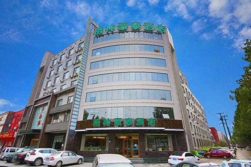 现代大楼，前有绿色招牌，周围是其他建筑，天空晴朗蔚蓝，充满各种物体，如停在大楼前的车辆、背景中的树木和街灯。整体氛围为繁忙的城市景象，人们可能在日常活动。
