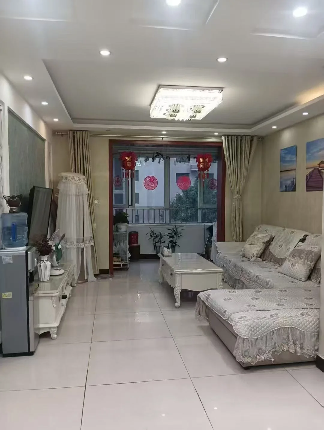 图片展示了一个宽敞的客厅，配有一张大白色的沙发和一张茶几。墙上安装了一个平板电视，地板上摆放着一盆盆栽。房间具有现代而优雅的氛围，通过窗户可以看到一个阳台。家具的摆放和装饰暗示着一个舒适和令人愉悦的放松和娱乐空间。