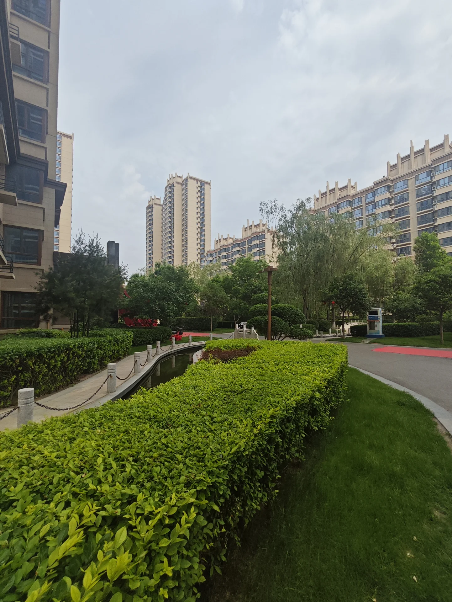 绿意盎然的公园，修剪整齐的灌木和草地，远处是高大的公寓和办公楼。阴郁的天空营造出宁静的氛围，公园为繁忙的城市环境提供了一个和平的避风港。绿树成荫和整洁的外观表明该地区受到居民和游客的喜爱。关键字:绿意盎然 修剪整齐 灌木 草地 高大建筑 公寓 办公楼 阴郁天空 宁静氛围 和平避风港