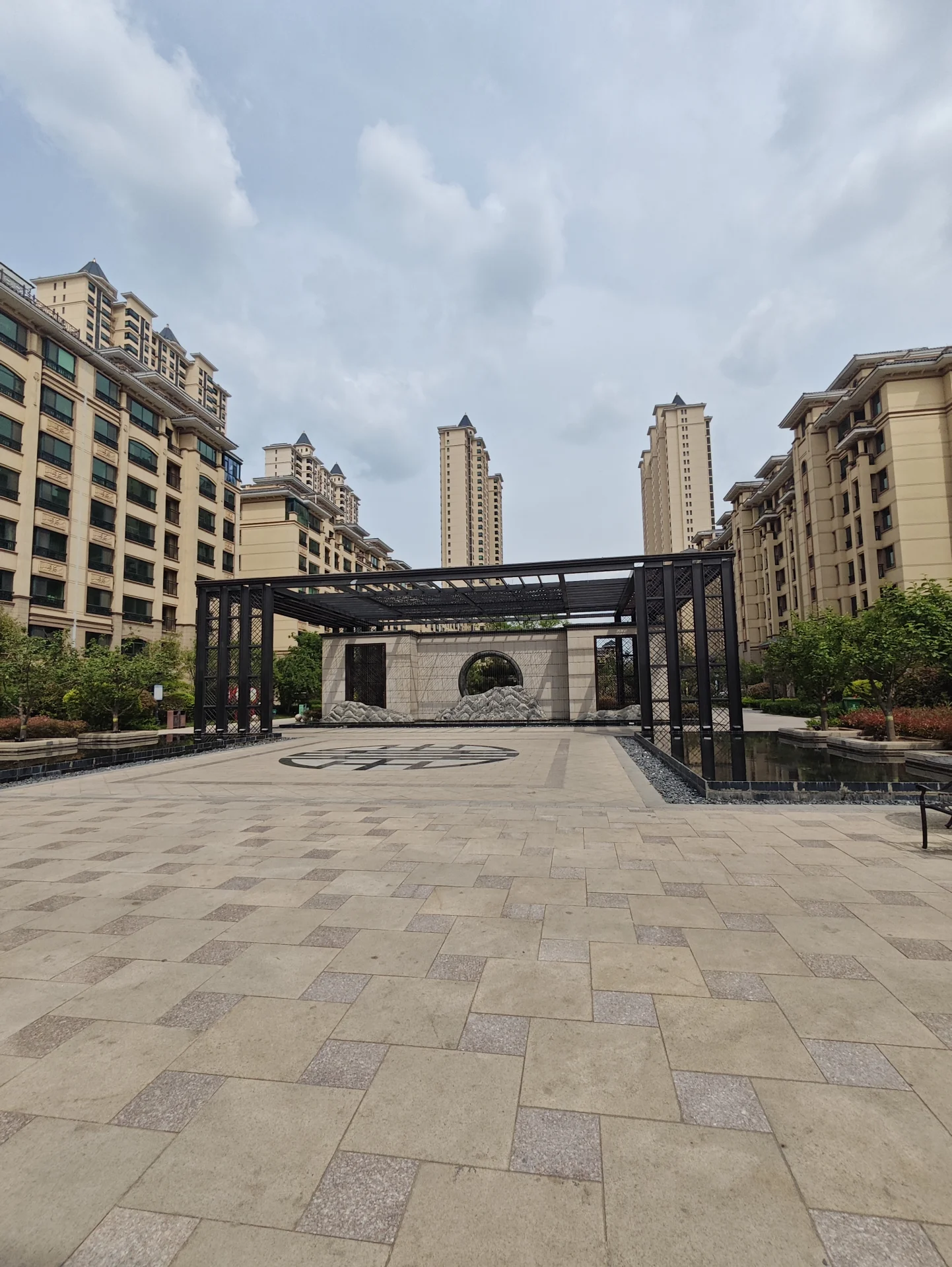 城市中心的开阔庭院，被高楼环绕，融合多种建筑风格。上方的伞为人们提供遮阴，营造出宁静而宜人的氛围。树木和植物增添自然气息，成为城市中的重要聚会场所，为繁忙街头提供一片宁静。关键字:开阔庭院 城市中心 高楼环绕 建筑风格 遮阴伞 宁静氛围 自然元素 聚会场所