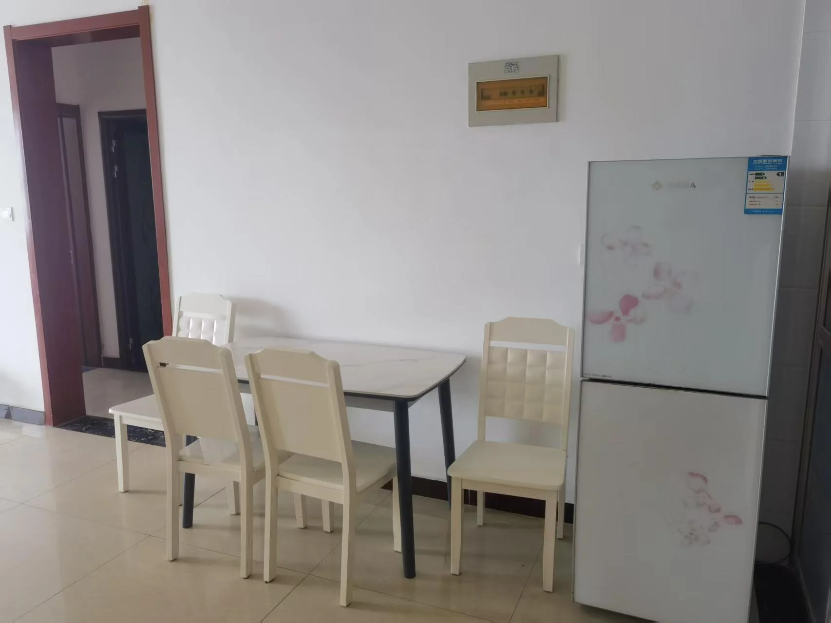 宽敞明亮的餐厅，白色家具搭配，餐桌四椅错落有致，冰箱背景增添厨房氛围，装饰画点缀墙面，钟表高悬餐桌上方，花瓶增添温馨气息。关键字:餐厅 白色家具 餐桌 椅子 冰箱 厨房氛围 装饰画 钟表 花瓶