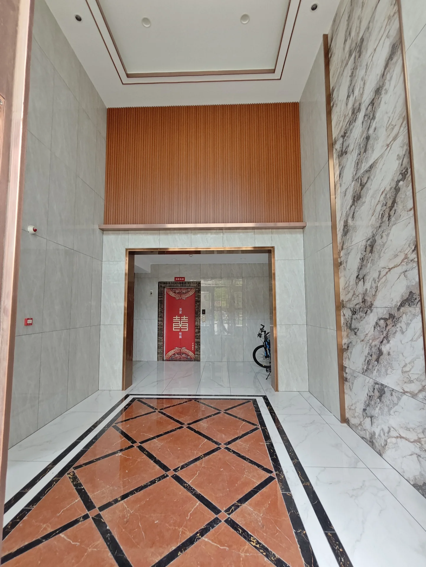 宽敞明亮的房间，大理石地面和墙面，中央红色门可能是入口或出口，门口停放着自行车，墙上挂有一只钟表增添实用性。整体氛围优雅而温馨。关键字:宽敞明亮 大理石地面 大理石墙面 红色门 自行车 钟表