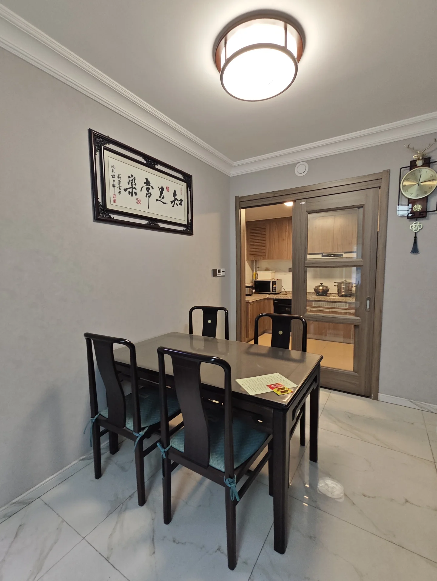简约现代餐厅，白色长桌配四把黑色椅子，桌上铺白桌布，菜单放置其上。房间以简洁明快为主，大窗户引入自然光线。墙上挂钟、桌上放书、窗台摆花瓶。氛围宁静高雅，餐厅成为空间焦点。