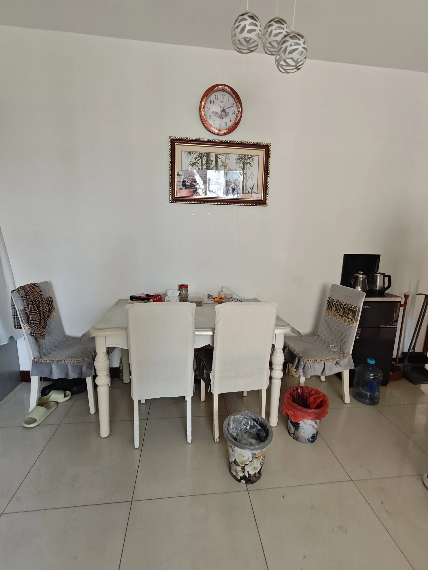 小而温馨的餐厅，白色墙面，木质桌椅，墙上挂着钟表。角落里摆放着一盆绿植，桌上散落着杯子、碗和瓶子，旁边有两尊花瓶。关键字:小餐厅 白色墙面 木质桌椅 钟表 绿植 杯子 碗 瓶子 花瓶