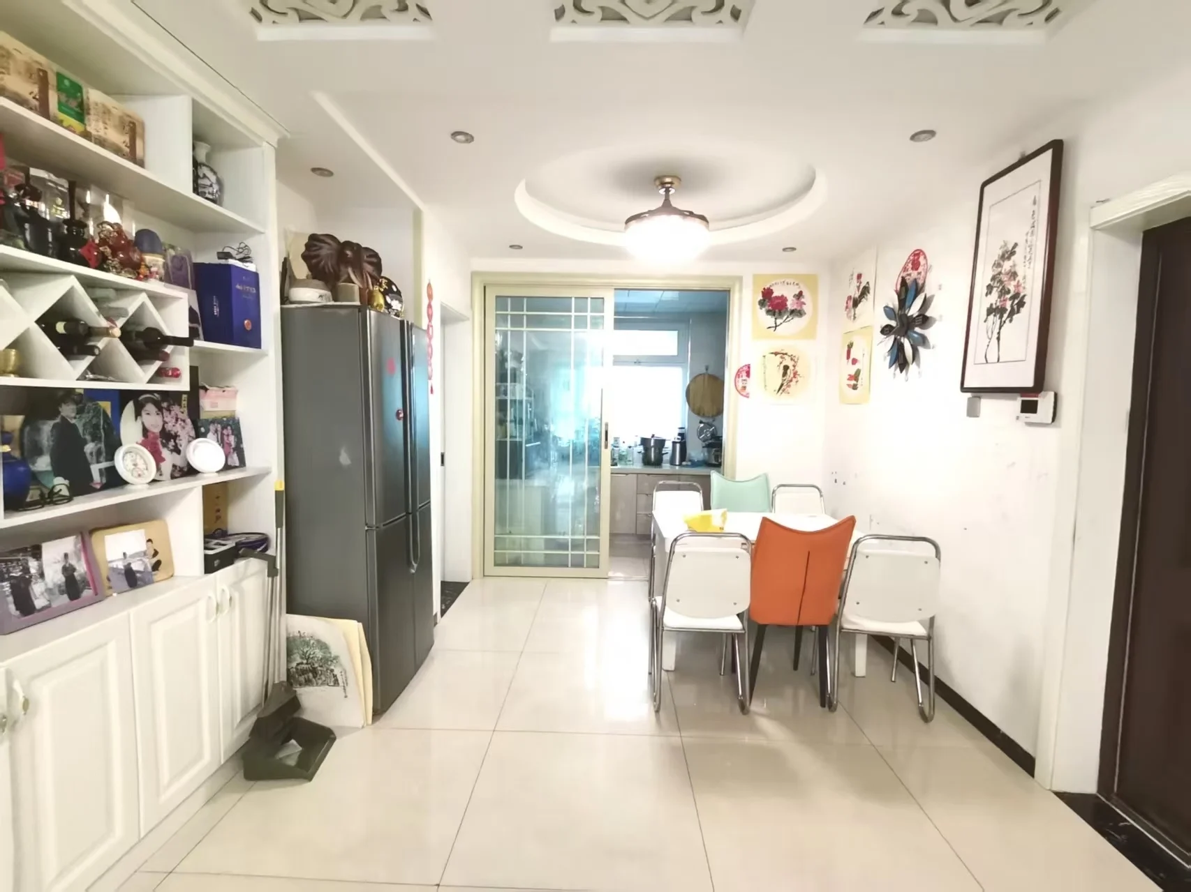 图片中展示了一个大的、开放且光线充足的餐厅，采用白色色调。餐厅设有长餐桌，周围摆放着几把椅子，营造出舒适友好的用餐氛围。房间内还设有厨房区域，配有冰箱和微波炉，暗示这是结合生活和用餐空间。房间内可见时钟、酒杯、杯子等物品，以及散落的书籍，显示出居住者喜欢阅读或拥有书籍收藏。这些物品和整体布局呈现出温馨且实用的居住氛围。关键字:餐厅, 厨房, 白色, 长餐桌, 椅子, 冰箱, 微波炉, 时钟, 酒杯, 书籍
