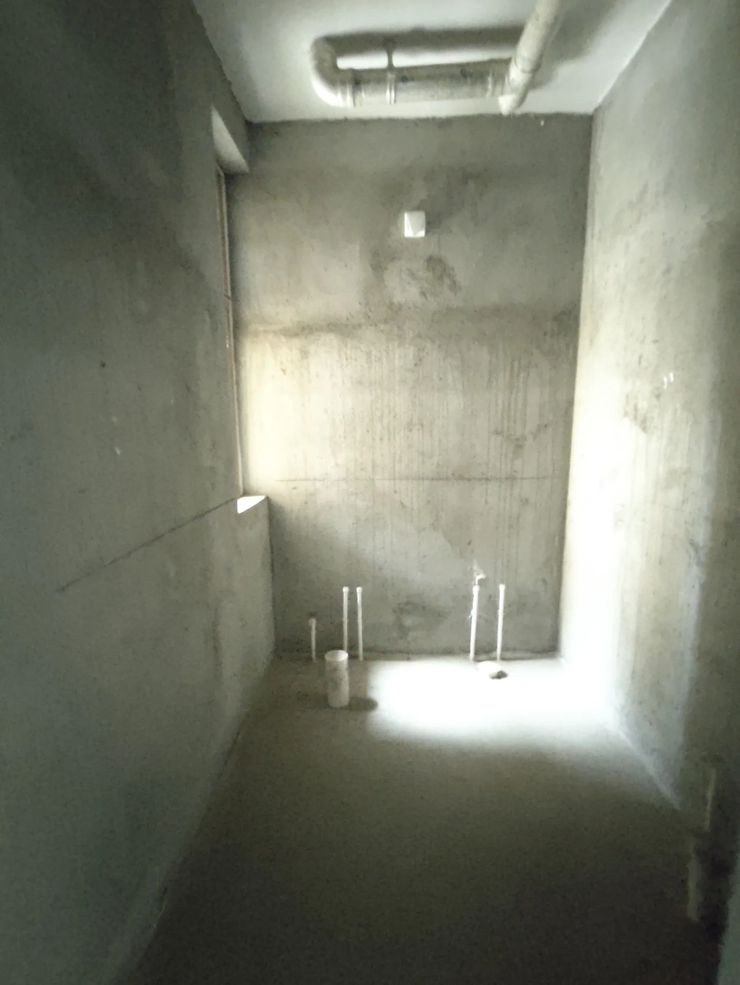 这间房间有混凝土墙和地板，中间摆放着一间看似在施工或未完成的厕所。一侧可以看到一根管道，房间内有两个窗户，自然光线洒进室内。关键字:混凝土墙 地板 厕所 施工 管道 窗户 自然光