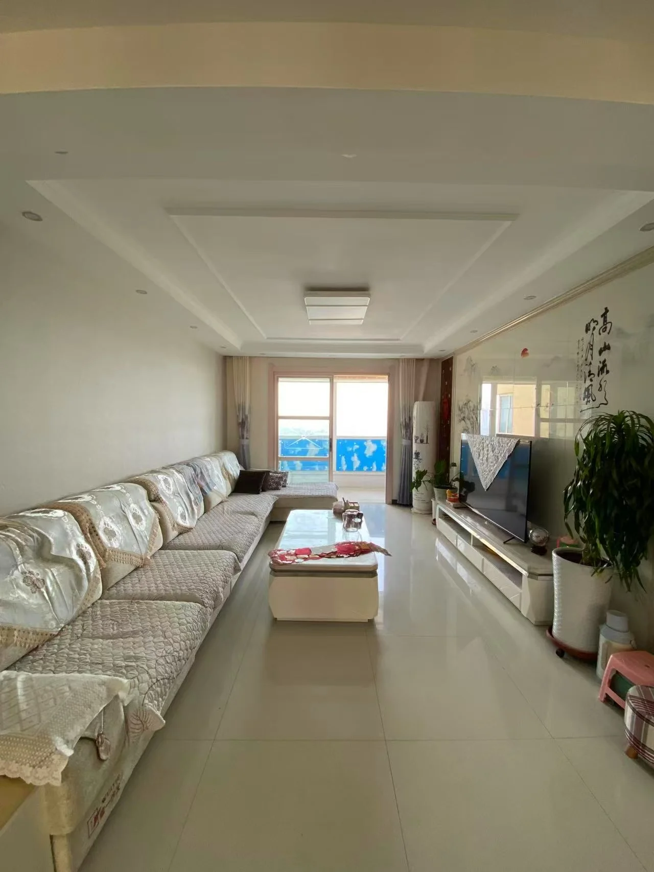 宽敞的客厅配有大型白色沙发和挂在墙上的平板电视。房间现代而优雅，天花板高且有大窗户，让自然光充满空间。客厅还配备了一张餐桌和几盆植物，为房间增添了一抹绿意和温暖。整体营造出一个舒适的休闲娱乐空间。关键字:客厅、白色沙发、平板电视、大窗户、自然光、餐桌、植物