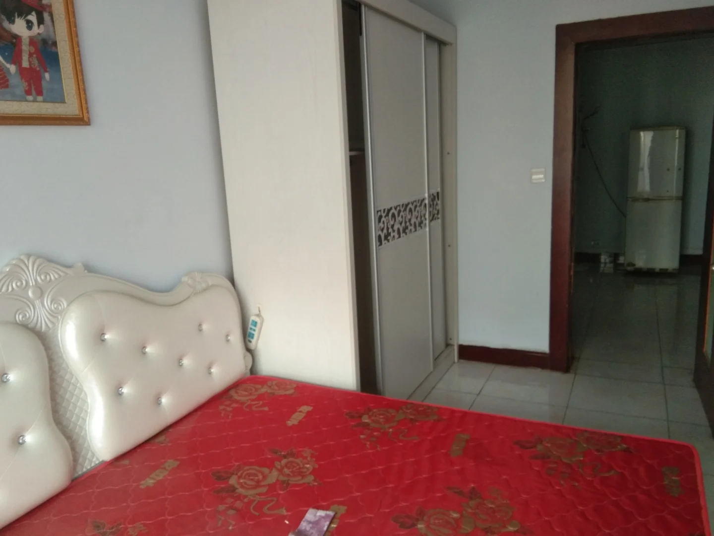 The image shows a bedroom with a white bed and red sheets. There is also a table in the room, which has a red cloth on it. A doorway can be seen next to the bed, leading into another room. The walls are painted blue, creating a calm atmosphere. A refrigerator is visible in the background, indicating 此处省略了... 多少字来着? .. 忘了...关键字: