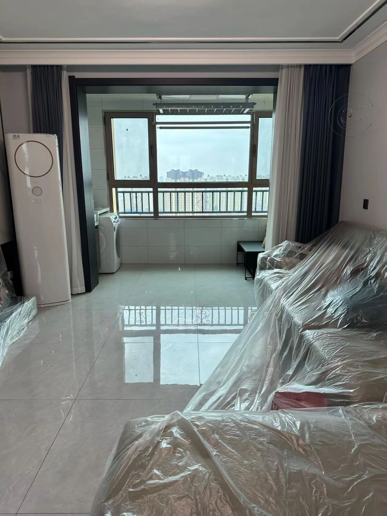 客厅空间，地面铺设大尺寸浅色瓷砖，配有白色壁挂式空调和部分家具（如沙发）被塑料布覆盖。采光良好，窗外可见城市景观，整体呈现现代简约风格。