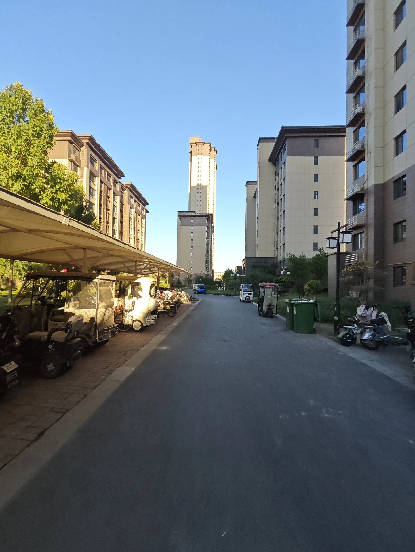 一条城市街道，两侧是多栋现代风格的住宅楼，街道中央有车辆和摩托车停放，背景天空晴朗。