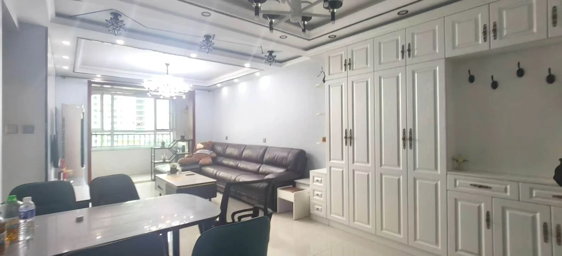 这是一个开放式空间，左侧设置了长条会议桌和椅子，右侧是大型白色储物柜，中间摆放着深色皮质沙发。整体装修风格偏向现代办公或多功能接待区，光线分布均匀。