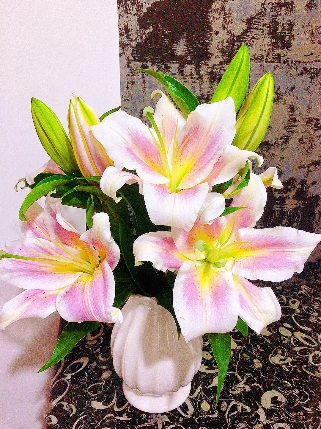 一束大型白色和粉色调的百合花插在白色花瓶中，背景为深色图案墙面，整体构图聚焦于鲜花本身。