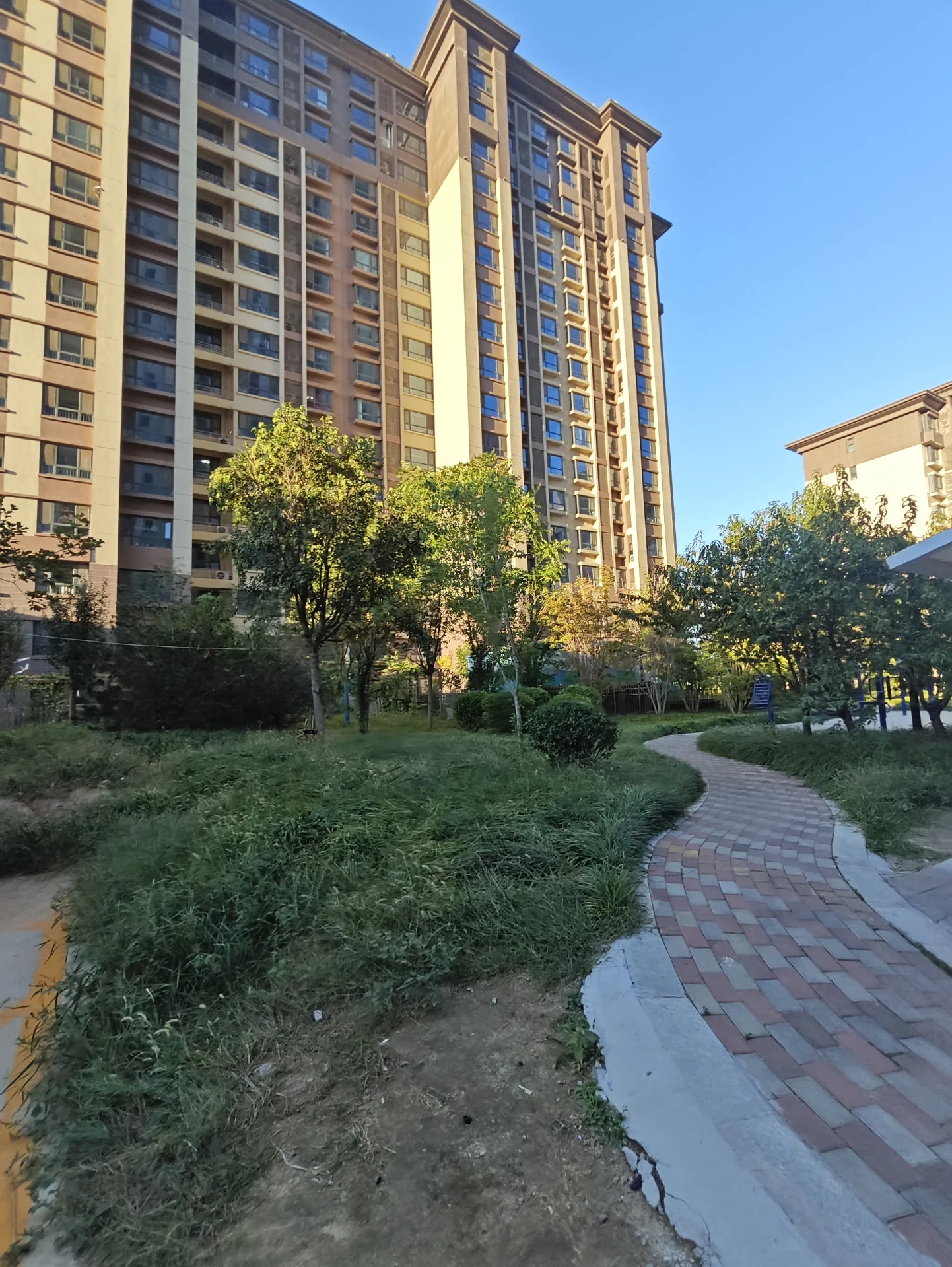 一座大型现代风格的住宅建筑，外立面呈米黄色调，结构宏大。建筑前方有修剪整齐的绿化带和蜿蜒的砖砌小径，整体环境宁静宜居。