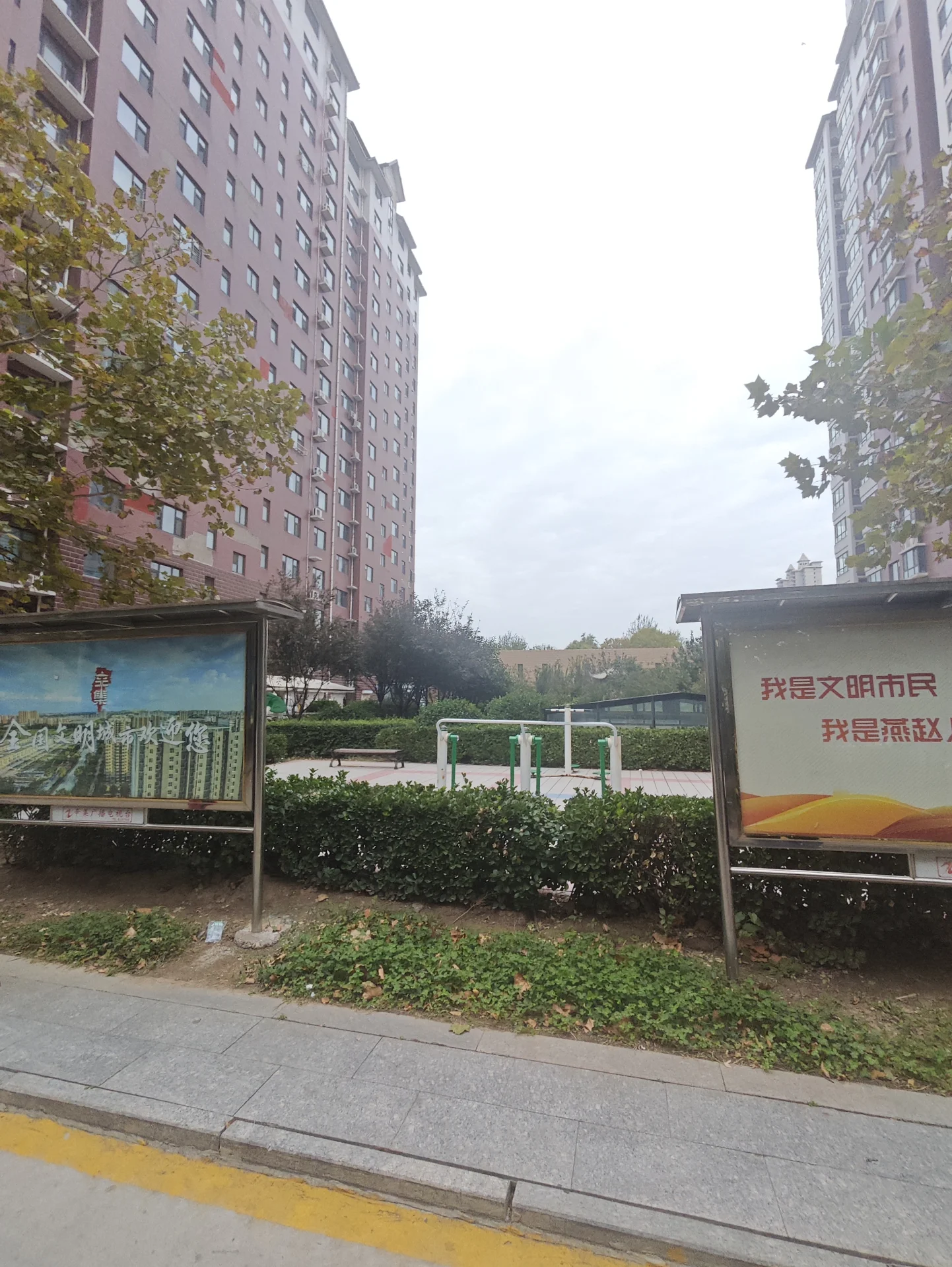 两栋高层住宅建筑矗立在街道两侧，中间有绿化带和儿童游乐设施。天空多云，整体呈现城市居住环境的日常景象。
