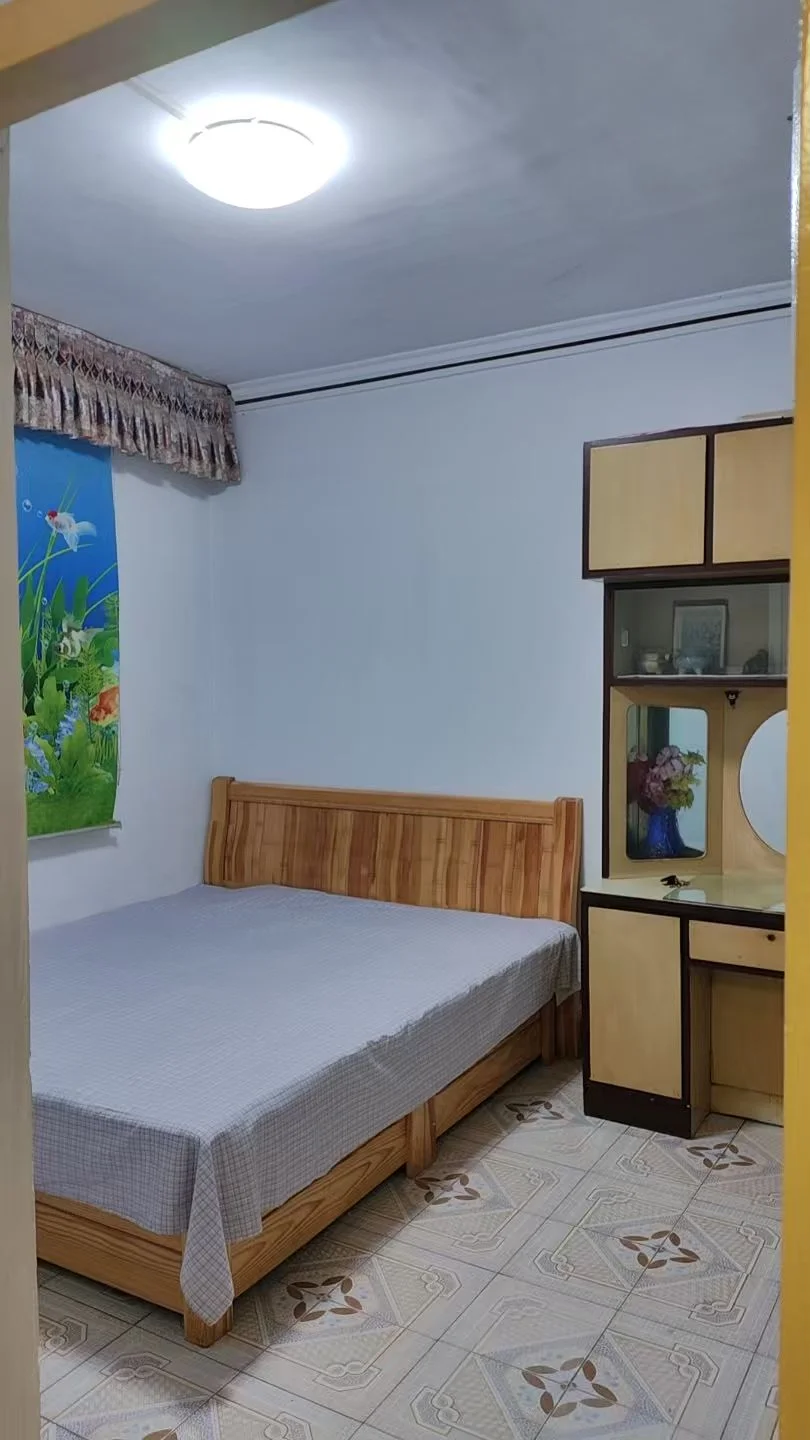 卧室空间，床为木质框架，搭配浅色床单。房间内光线充足，墙面简洁，整体装修风格偏向基础实用型。