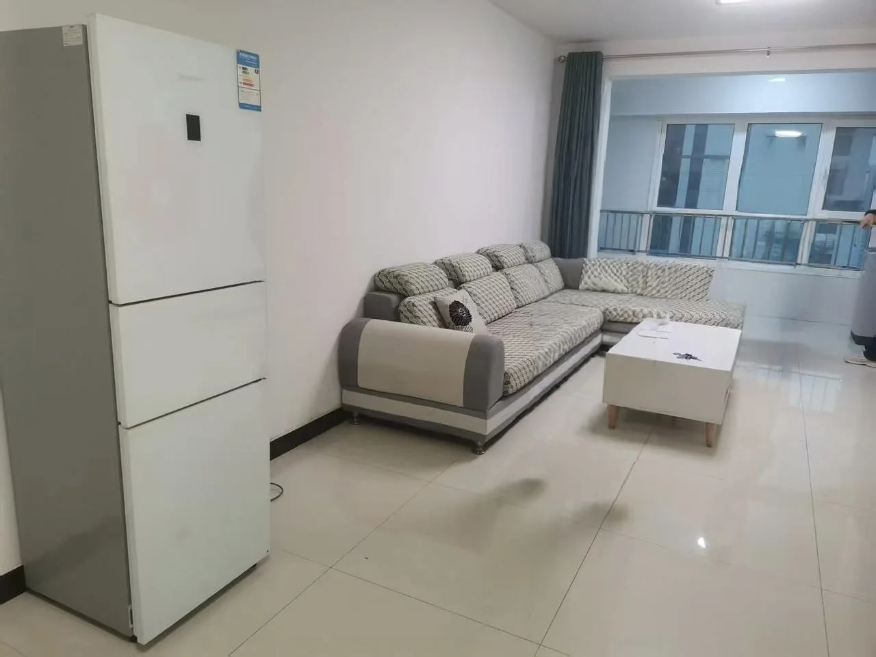 客厅空间，地面铺设浅色瓷砖，配有一套深灰色布艺沙发和白色茶几。房间采光良好，整体装修风格偏向现代简约，空间显得开阔明亮。