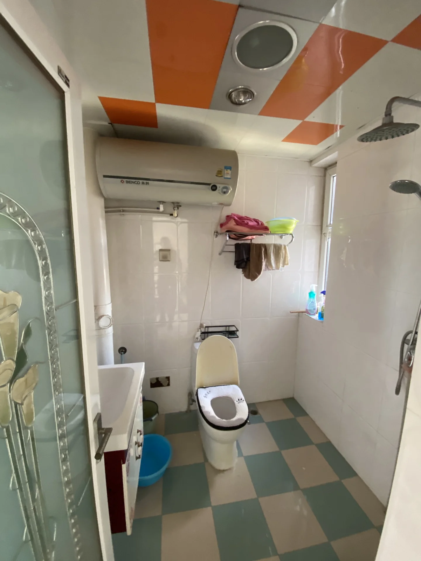 卫生间空间，墙面和地面采用不同色调的瓷砖铺设，天花板设计有几何图案装饰。房间内配备了吊顶空调、马桶和淋浴区域，整体环境功能明确。