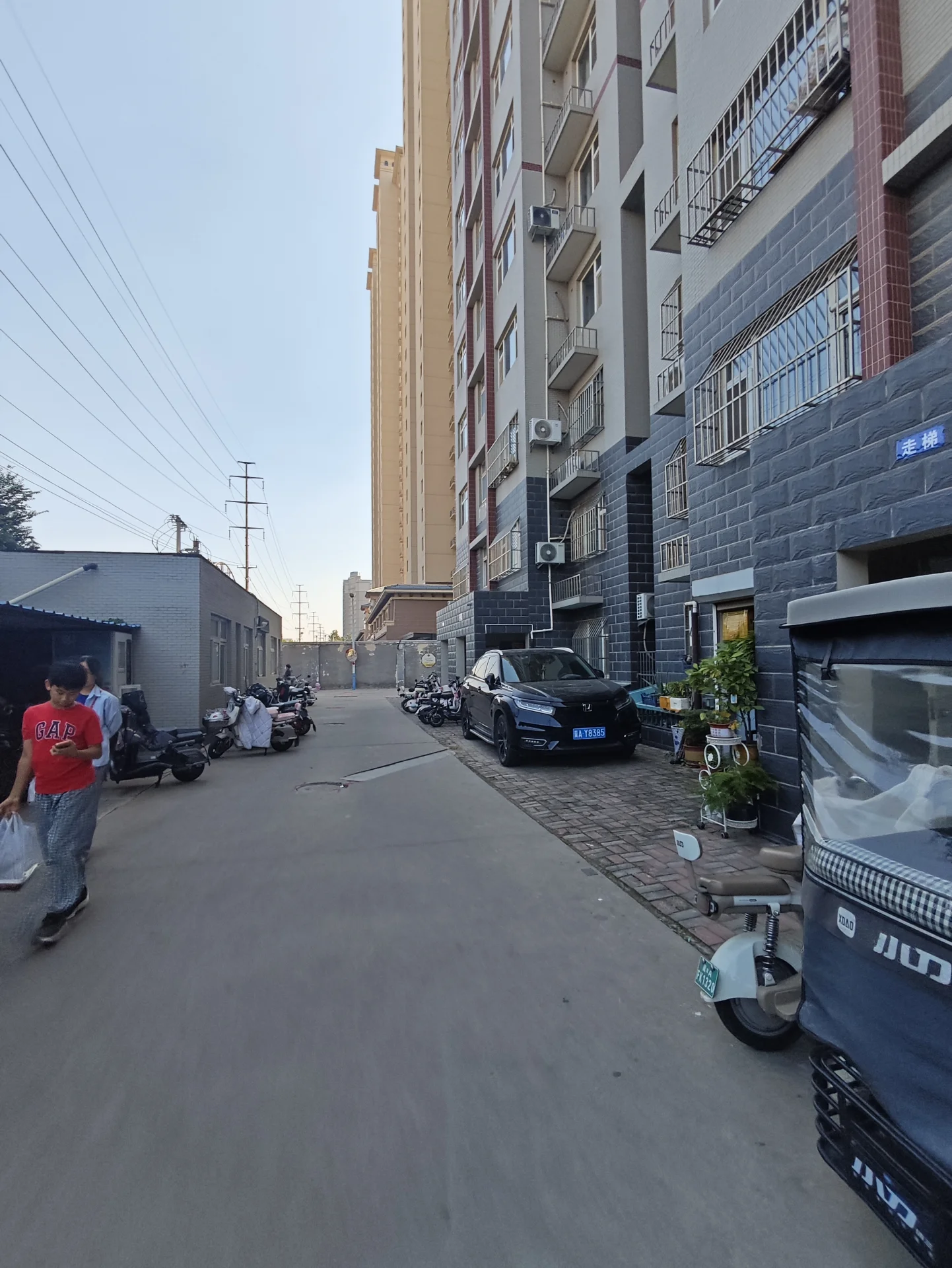 一条城市街道，两侧是不同风格的住宅建筑，背景中有一栋高层现代建筑。街道上停放着车辆和摩托车，整体呈现典型的城市居住环境。