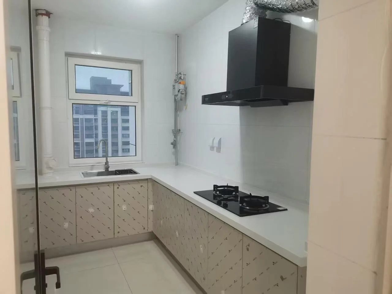 厨房空间，橱柜采用浅色纹理设计，配备不锈钢水槽和黑色抽油烟机。墙面和地面铺设浅色瓷砖，采光良好，整体呈现现代简约的装修风格。