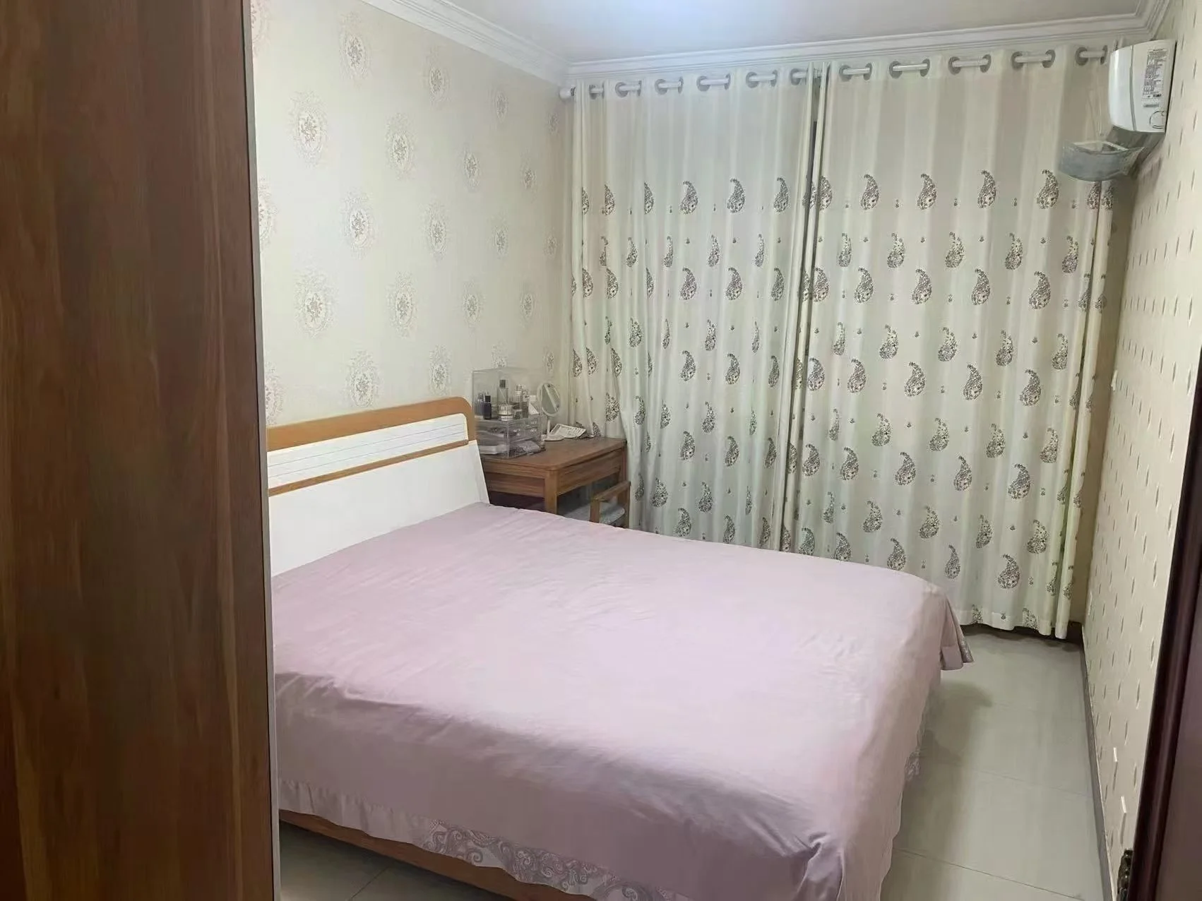 卧室空间，床铺为木质框架，搭配粉色床品。房间内采光适中，墙面有装饰性壁纸，整体风格偏向温馨和基础实用型。