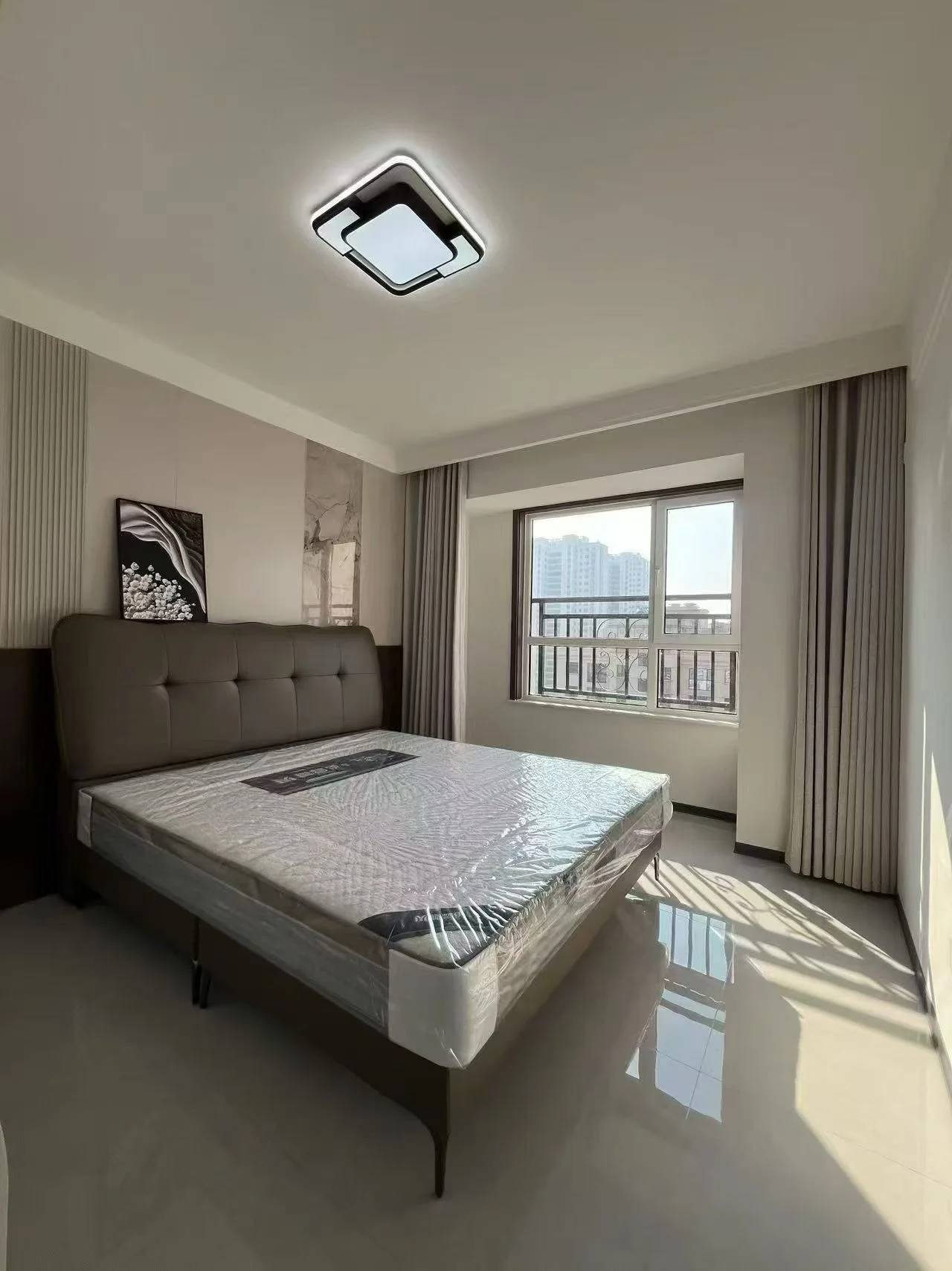 卧室空间，床铺为深色软包材质，搭配白色床品。房间采光良好，通过大窗引入自然光，整体装修风格偏向现代简约，环境整洁舒适。