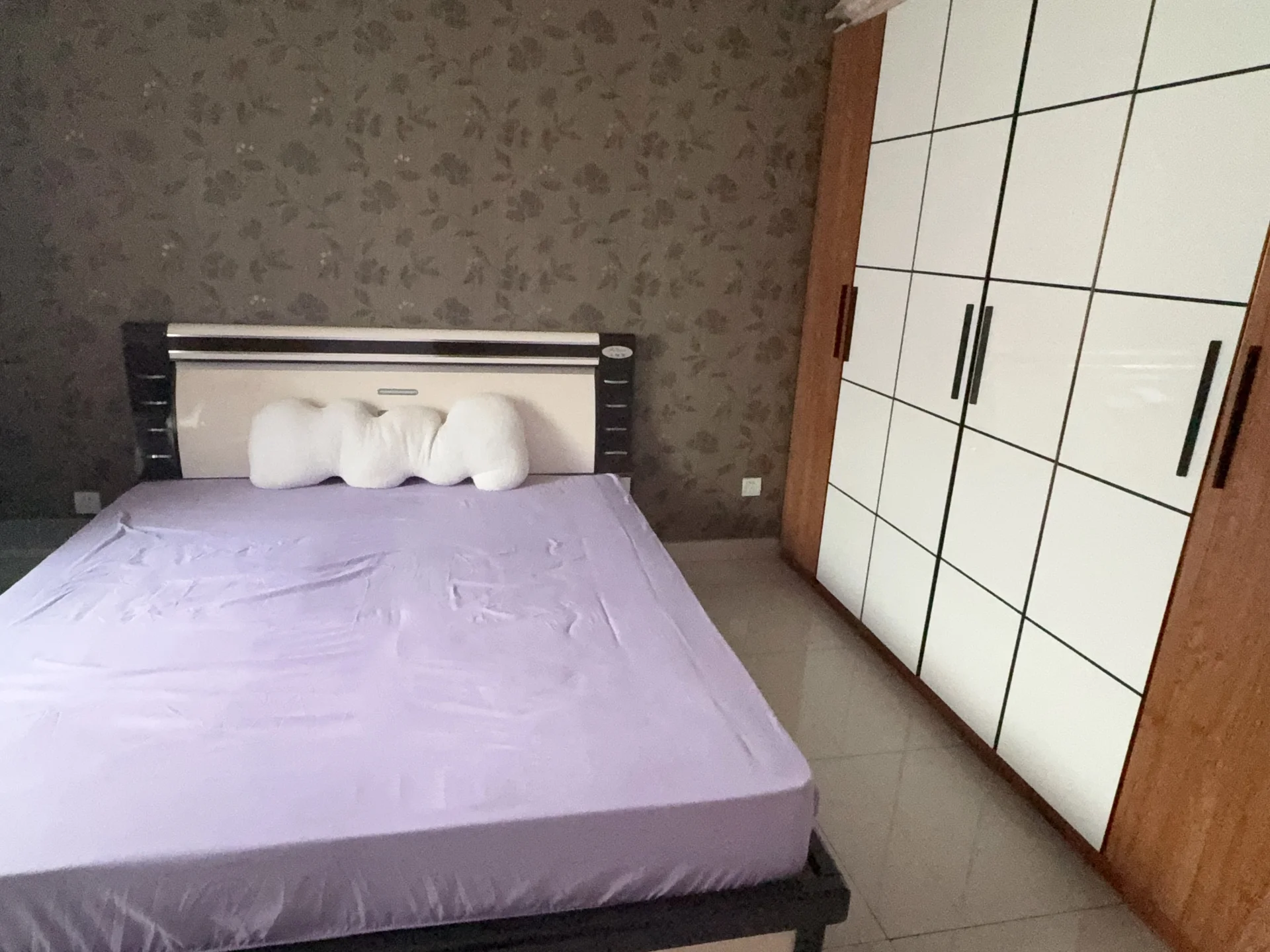 卧室场景，床铺为深色框架搭配白色床品，背景墙采用灰白色图案装饰。房间光线适中，整体呈现基础且整洁的居住环境。