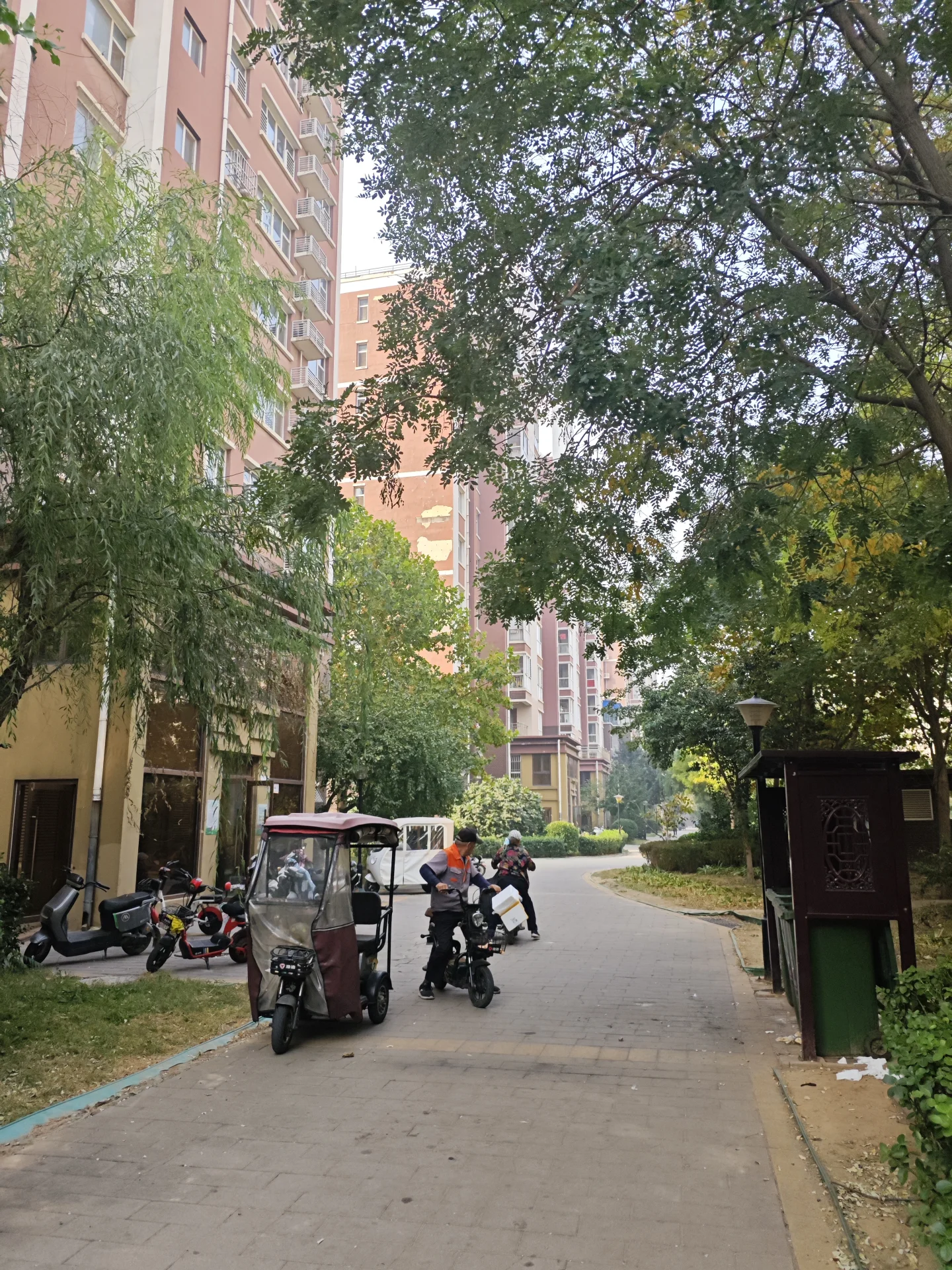 一条绿树环绕的社区内部道路，两侧是现代风格的住宅楼，路边停放着几辆摩托车和一辆三轮车，整体环境绿化良好，氛围宁静宜居。