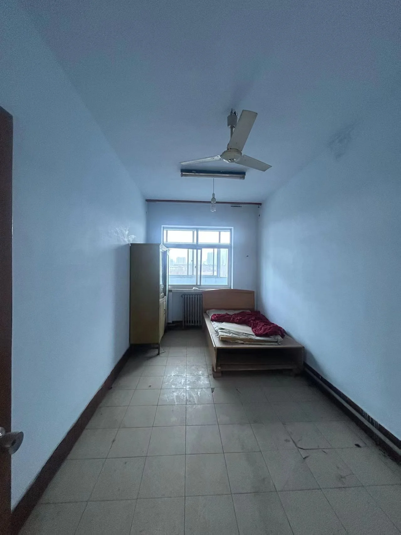 卧室空间，地面铺设浅色瓷砖，配有衣柜和一张床。房间内安装了吊扇和空调，采光适中，整体呈现基础实用风格。
