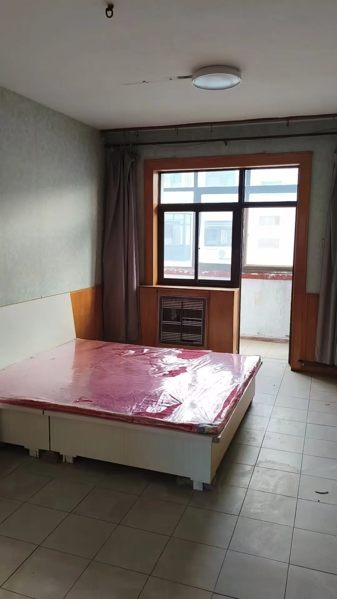 卧室空间，地面铺设浅色瓷砖，房间内摆放着一张覆盖塑料膜的床架。采光适中，整体环境简洁实用。