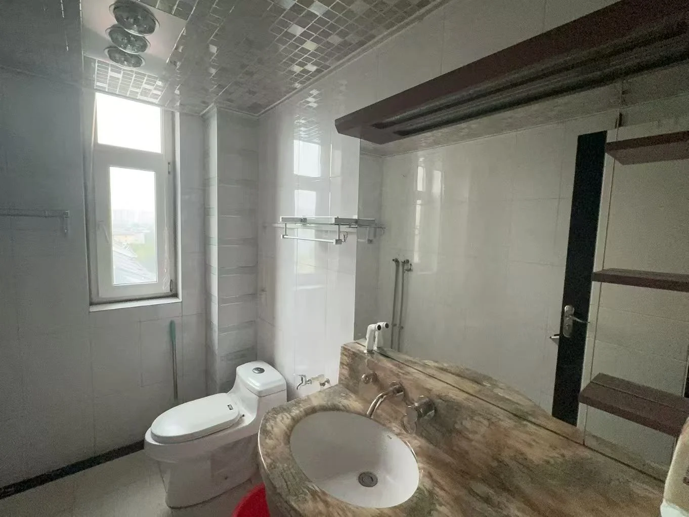 卫生间内部场景，墙面和地面铺设浅色瓷砖，配有白色马桶和石材台面洗手盆。空间采光良好，整体风格简洁实用。