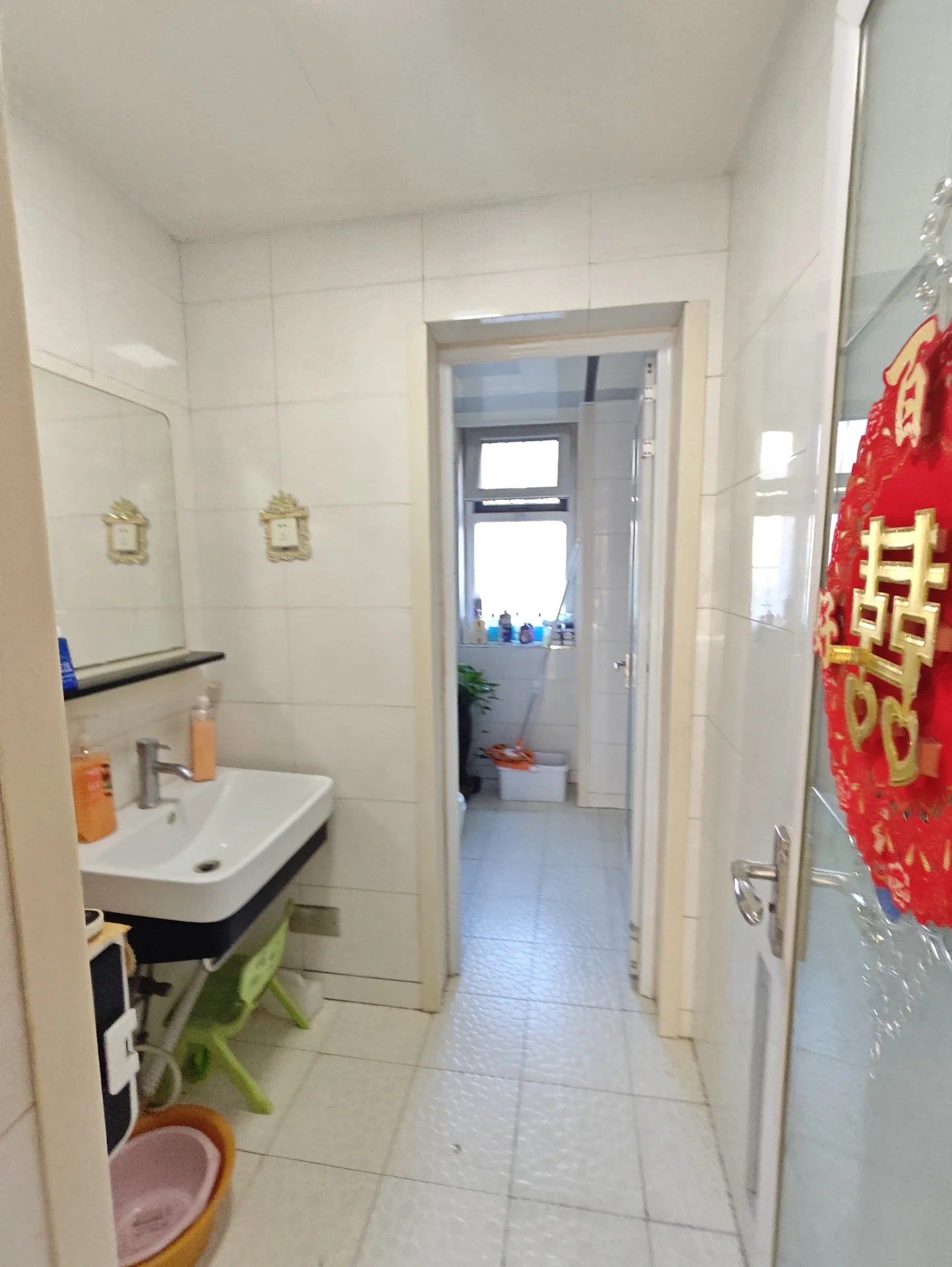 卫生间内部场景，墙面和地面均采用白色瓷砖铺设，设计简洁明亮。空间布局清晰，配备了白色洗手台和镜子，整体环境基础且干净。