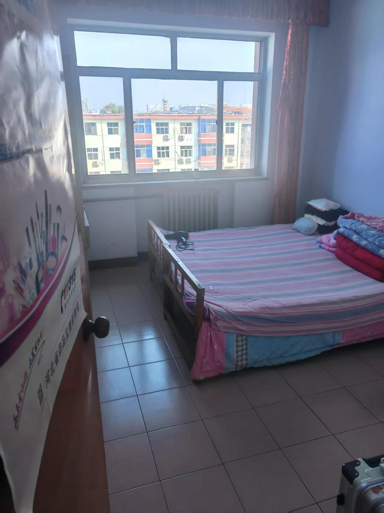 卧室空间，铺设深色瓷砖地面，配有单人床和窗帘。房间采光良好，整体装修风格偏基础实用型。