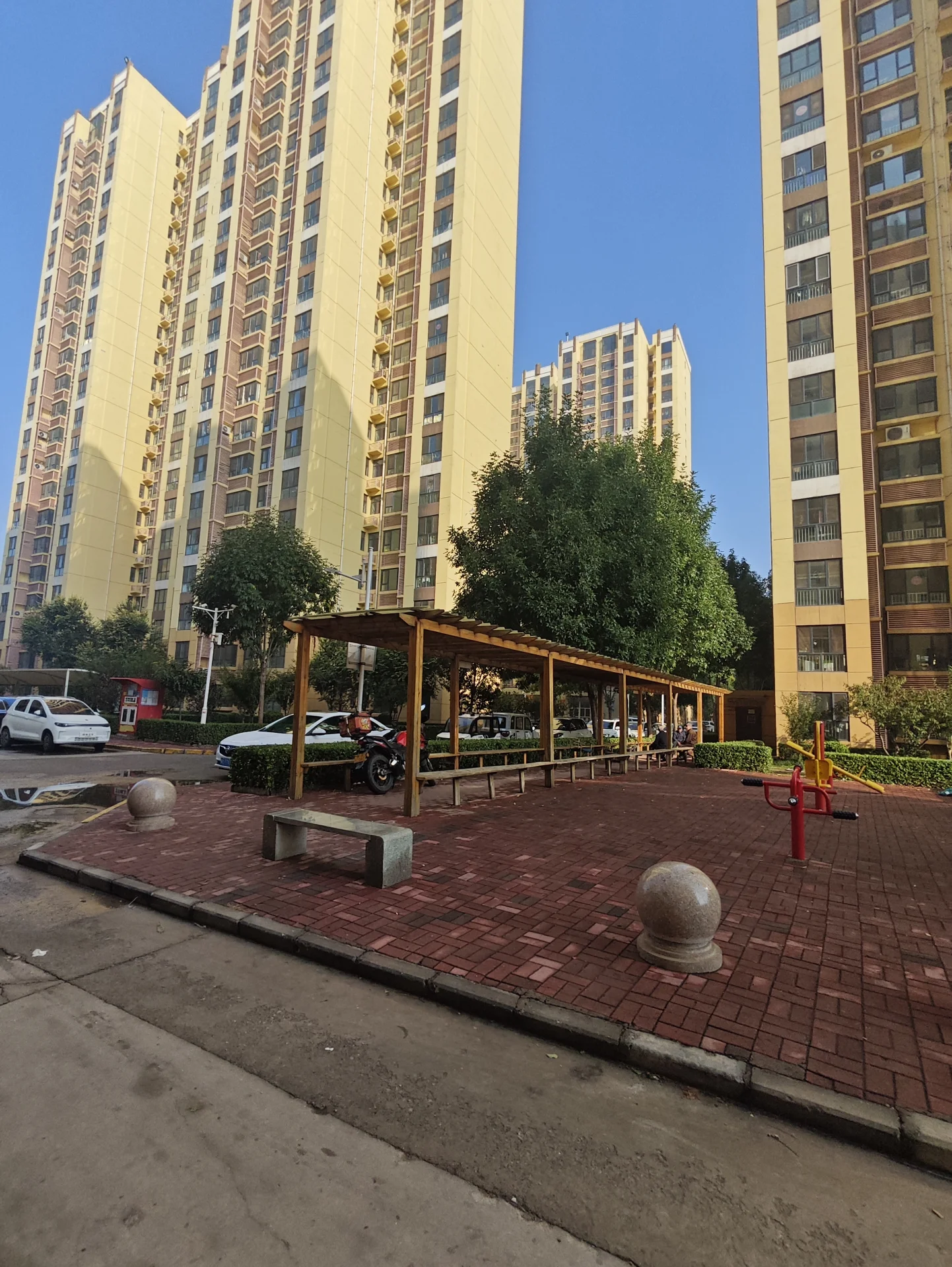 一个户外儿童游乐区，地面铺设红砖，配有木质凉亭和部分游乐设施。背景是高层住宅楼，整体环境在城市中具有一定的居住配套氛围。