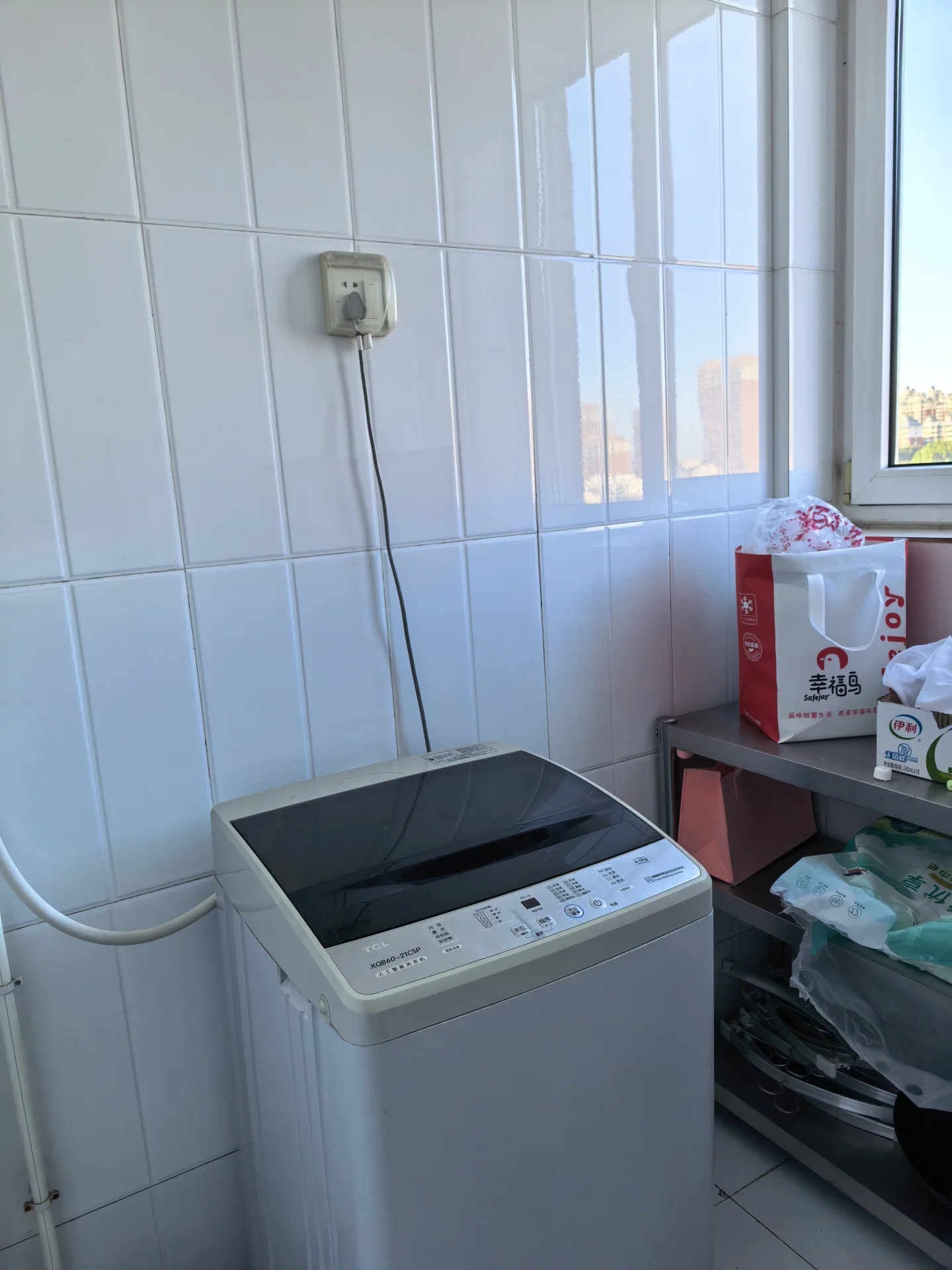 洗衣机放置在白色瓷砖墙前，背景可见窗外城市建筑的轮廓。场景属于功能性设备展示，整体环境简洁明亮。