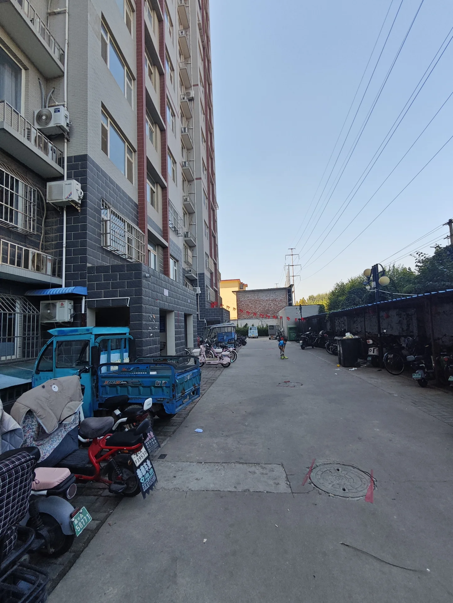 一栋高层住宅建筑占据画面主体，外立面采用浅色调砖石材质，周围停放着多辆摩托车和小型车辆。街道相对开阔，环境显示出城市居住区的日常景象。