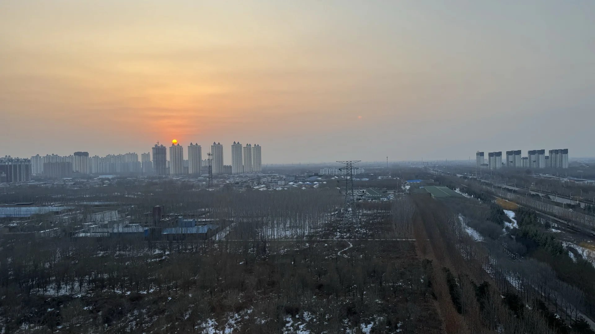 日出时，城市建筑群在远景中依稀可见，前景是广阔的未开发土地和稀疏的树林，整体呈现出一种开阔而宁静的城市边缘景象。