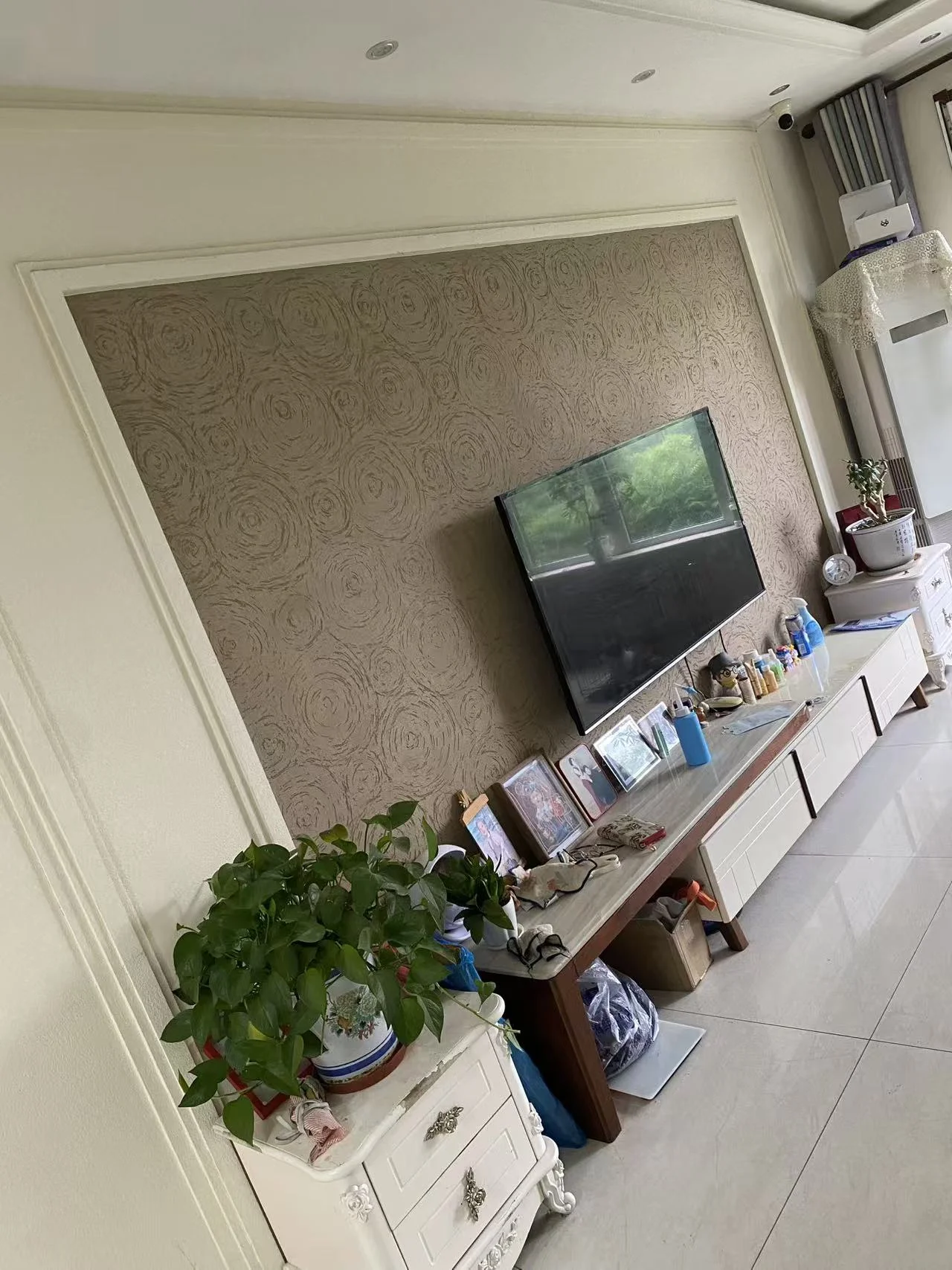 客厅场景，墙面采用纹理壁纸装饰，配有电视和储物柜。地面为浅色瓷砖，整体装修风格偏向现代简约，光线适中。