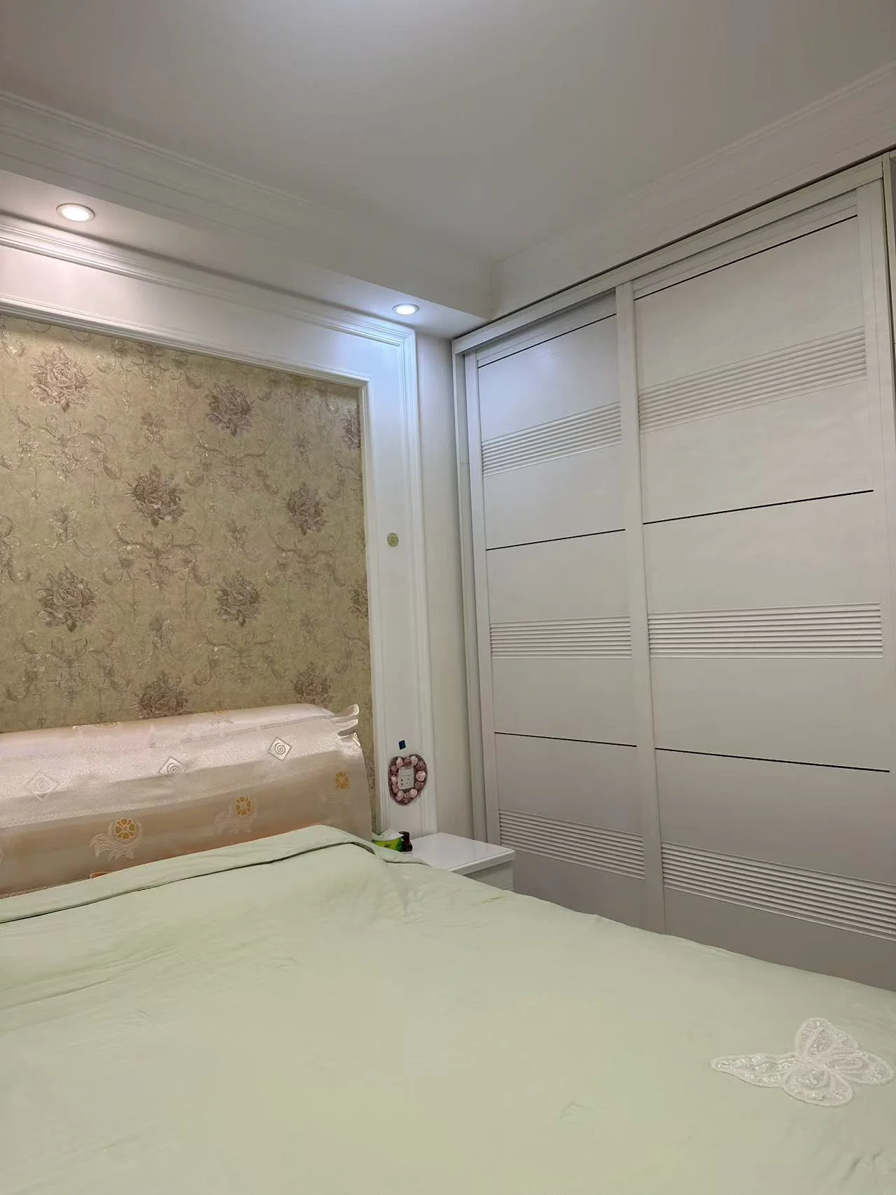 卧室空间，床铺采用浅绿色床品，背景墙装饰有淡雅的纹理图案。房间内配备了大型白色衣柜，整体采光良好，呈现出温馨舒适的居住氛围。