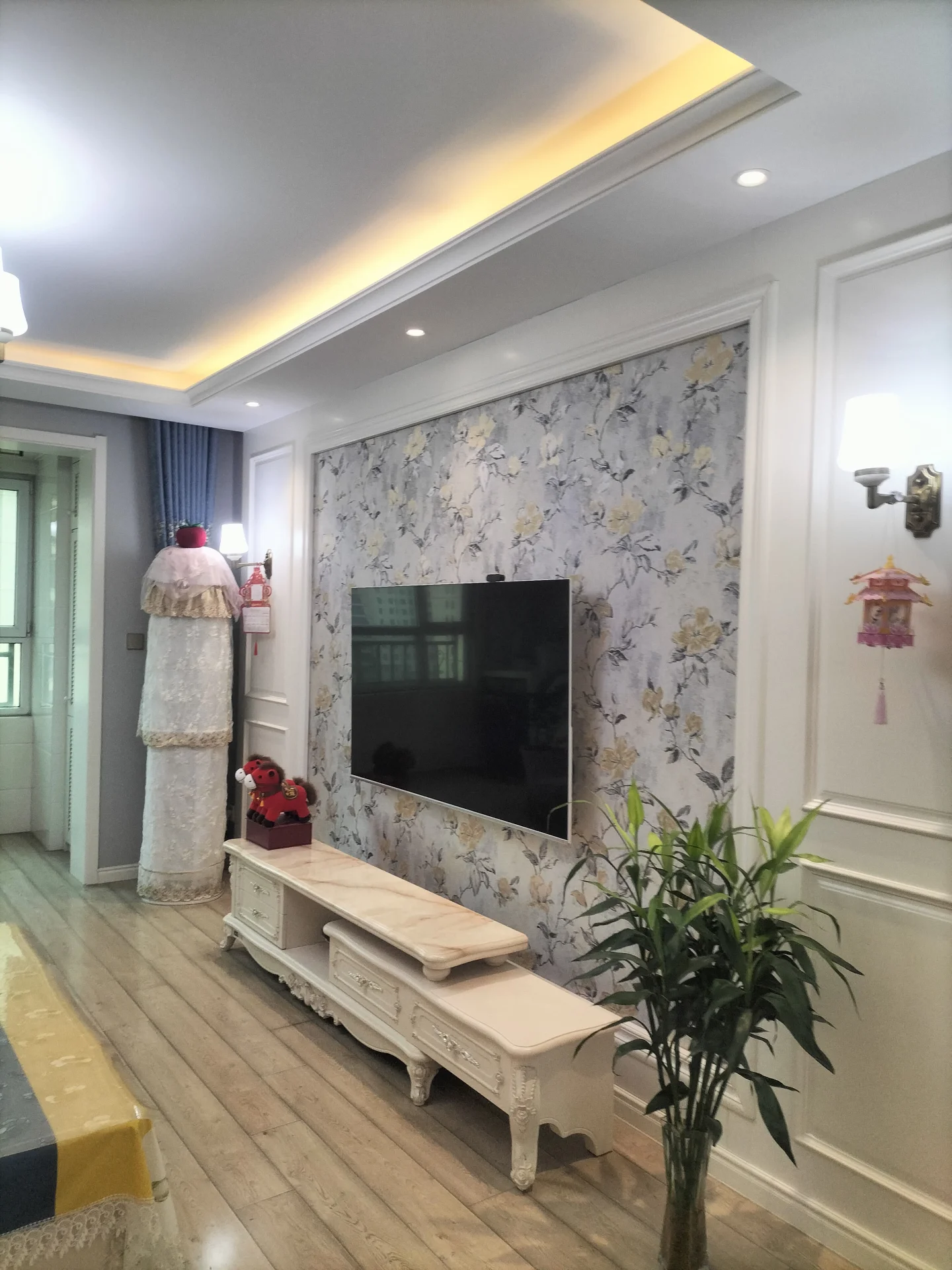 客厅场景，墙面采用淡雅的印花壁纸作为背景，配有白色雕花电视柜和大型平板电视。天花板设计有嵌入式灯带，采光均匀，整体呈现古典与现代结合的装修风格。