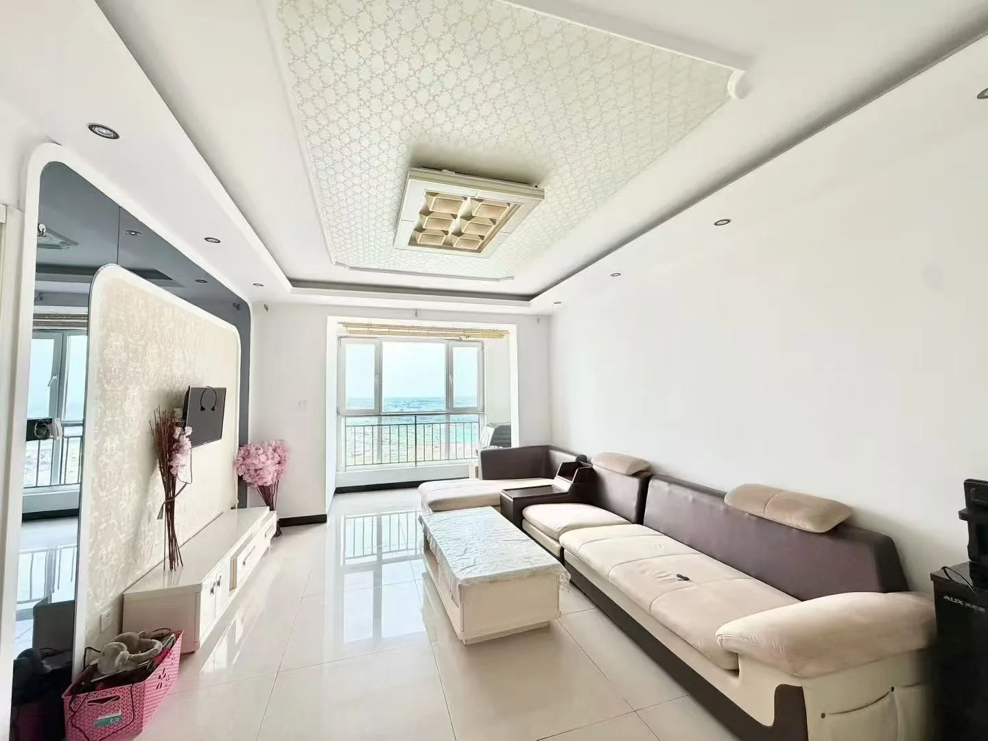 客厅空间，地面铺设大尺寸白色瓷砖，天花板采用几何纹理设计。房间内摆放有深色沙发和茶几，采光良好，整体呈现现代简约风格。